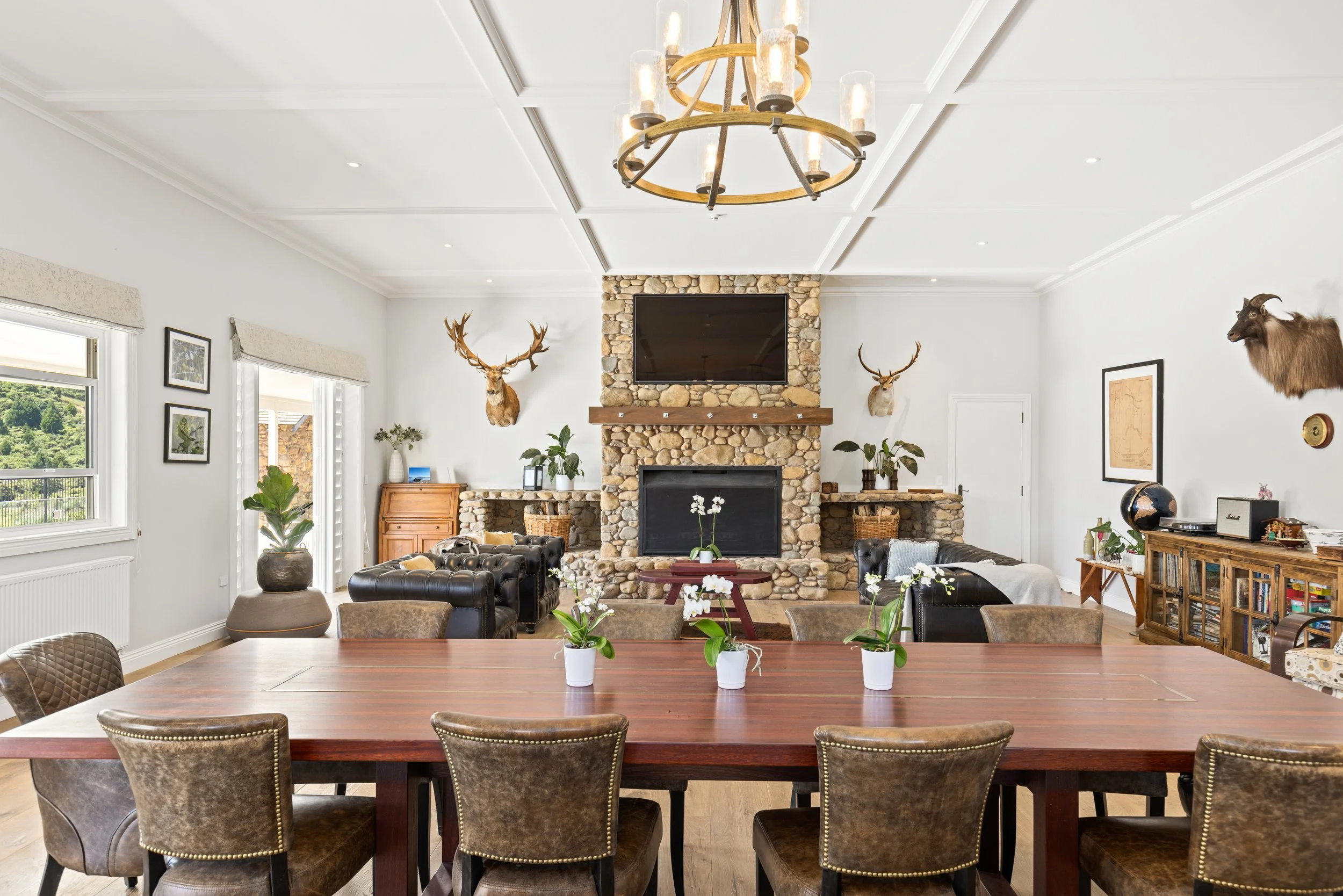 A living and dining room with a wooden dining table, seven chairs, potted plants, a stone fireplace with a mounted television and animal head trophies, windows with blinds, a chandelier, and various decor items.