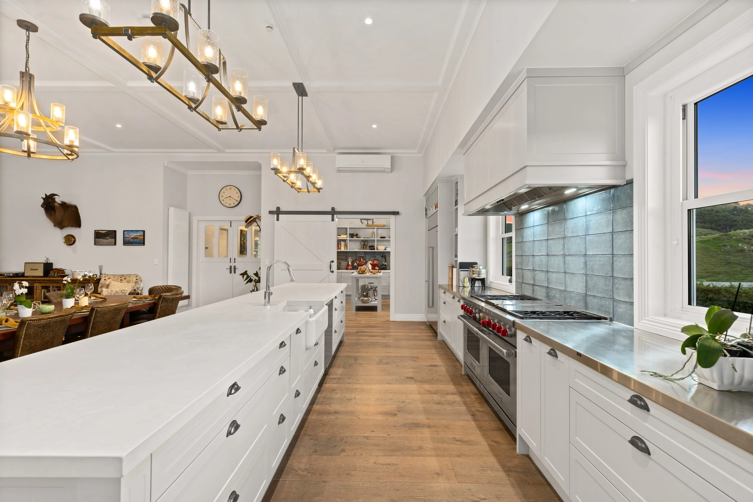 Modern white kitchen with large island, wooden floors, and view of sunset through window.