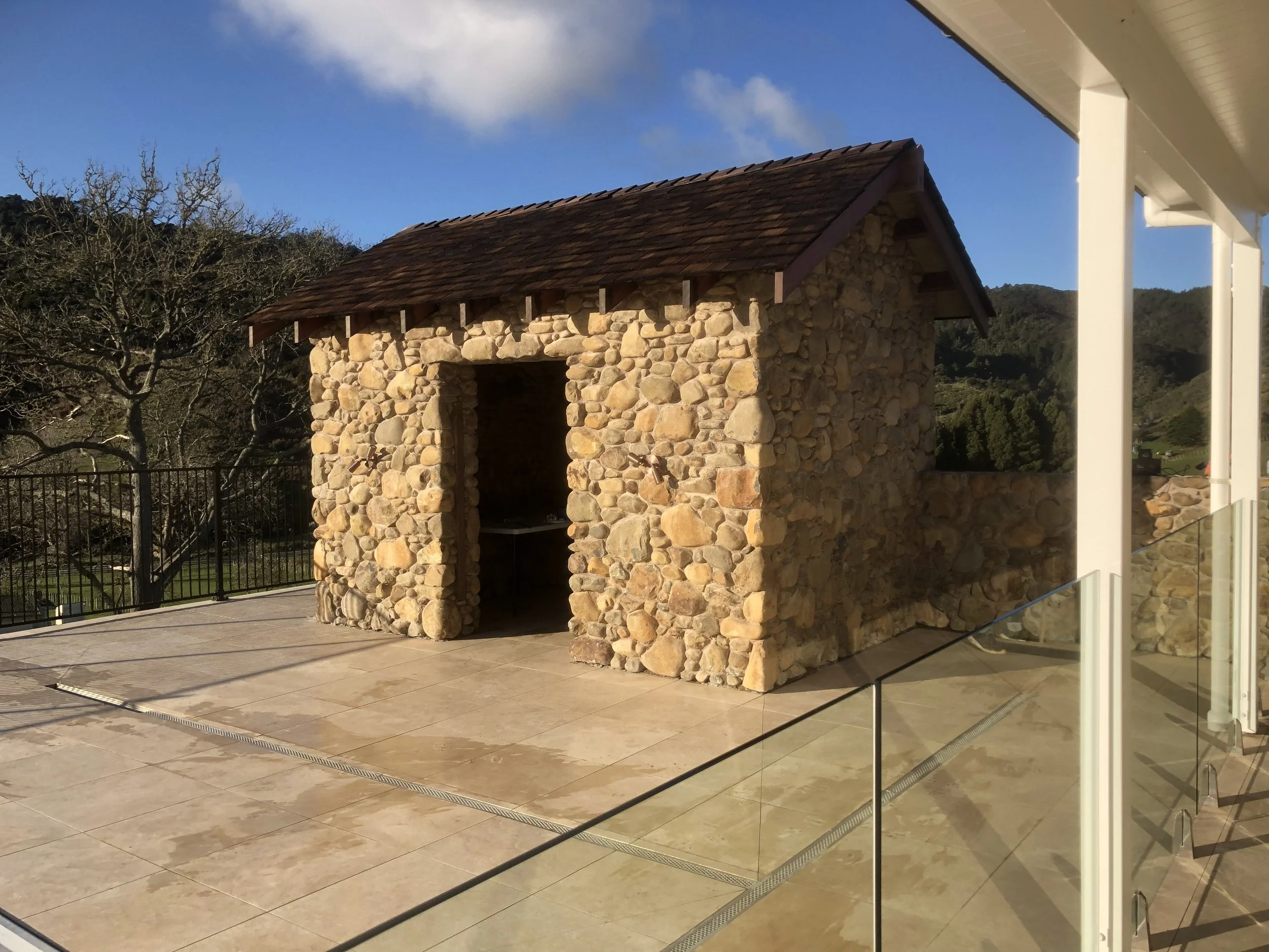 A stone building with a wooden shingle roof on a patio with beige tiles, surrounded by a metal fence and overlooking a hilly landscape with trees under a blue sky.