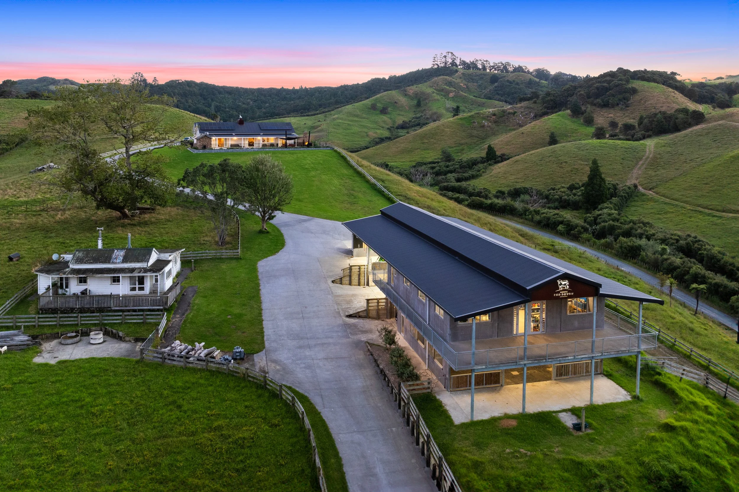 A rural landscape at sunset with hills, scattered trees, green fields, and a house with solar panels on the roof. There are also several buildings, including a barn and a house with a porch, and a driveway leading to the structures.