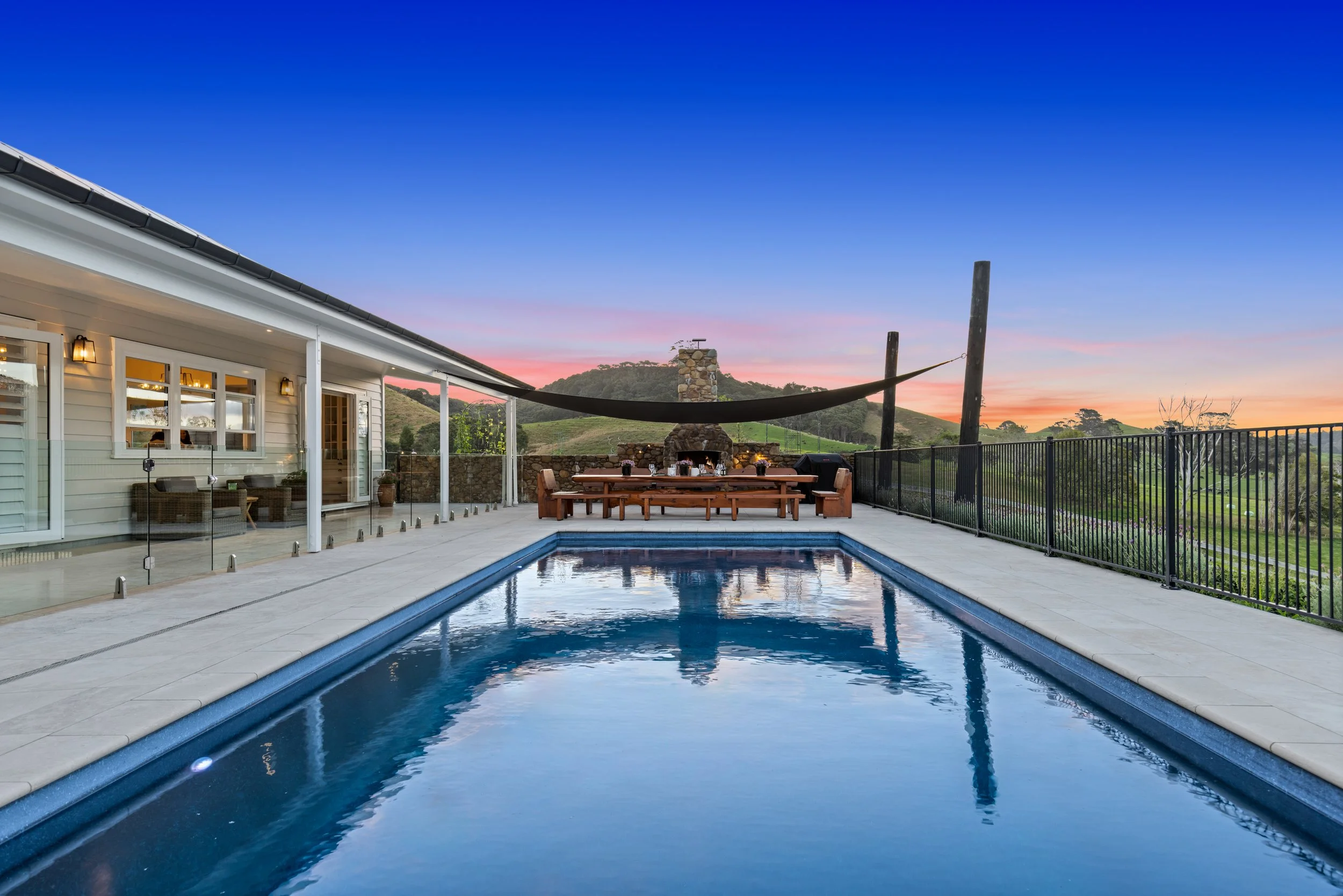 A backyard with a swimming pool, outdoor dining table, and fireplace at sunset, hillside landscape in the background, and a house with lit porch lights on the left.