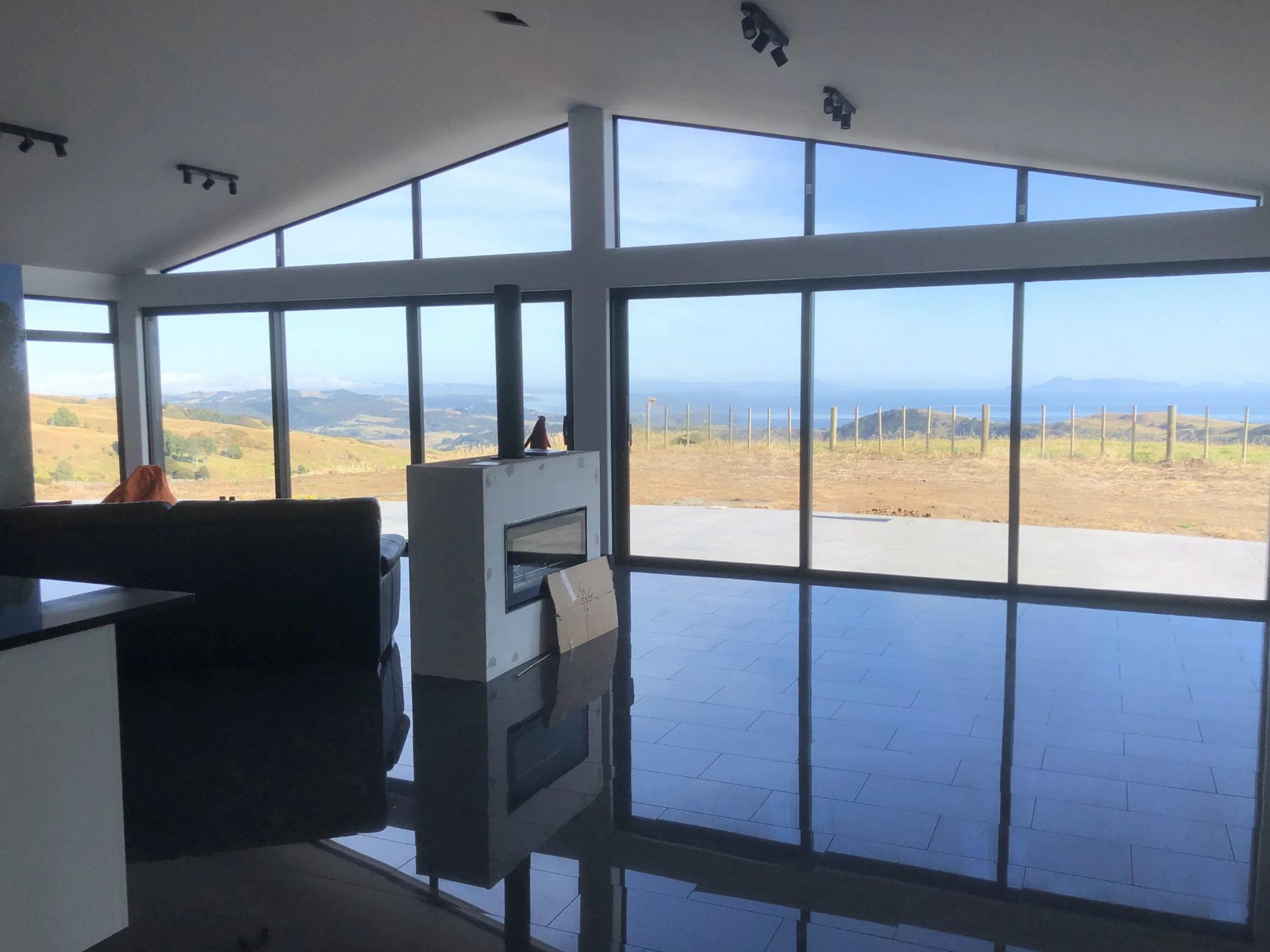 Modern living room with large floor-to-ceiling windows overlooking a landscape of hills, coastline, and ocean in the distance, with a white fireplace in the center and black sofas nearby.