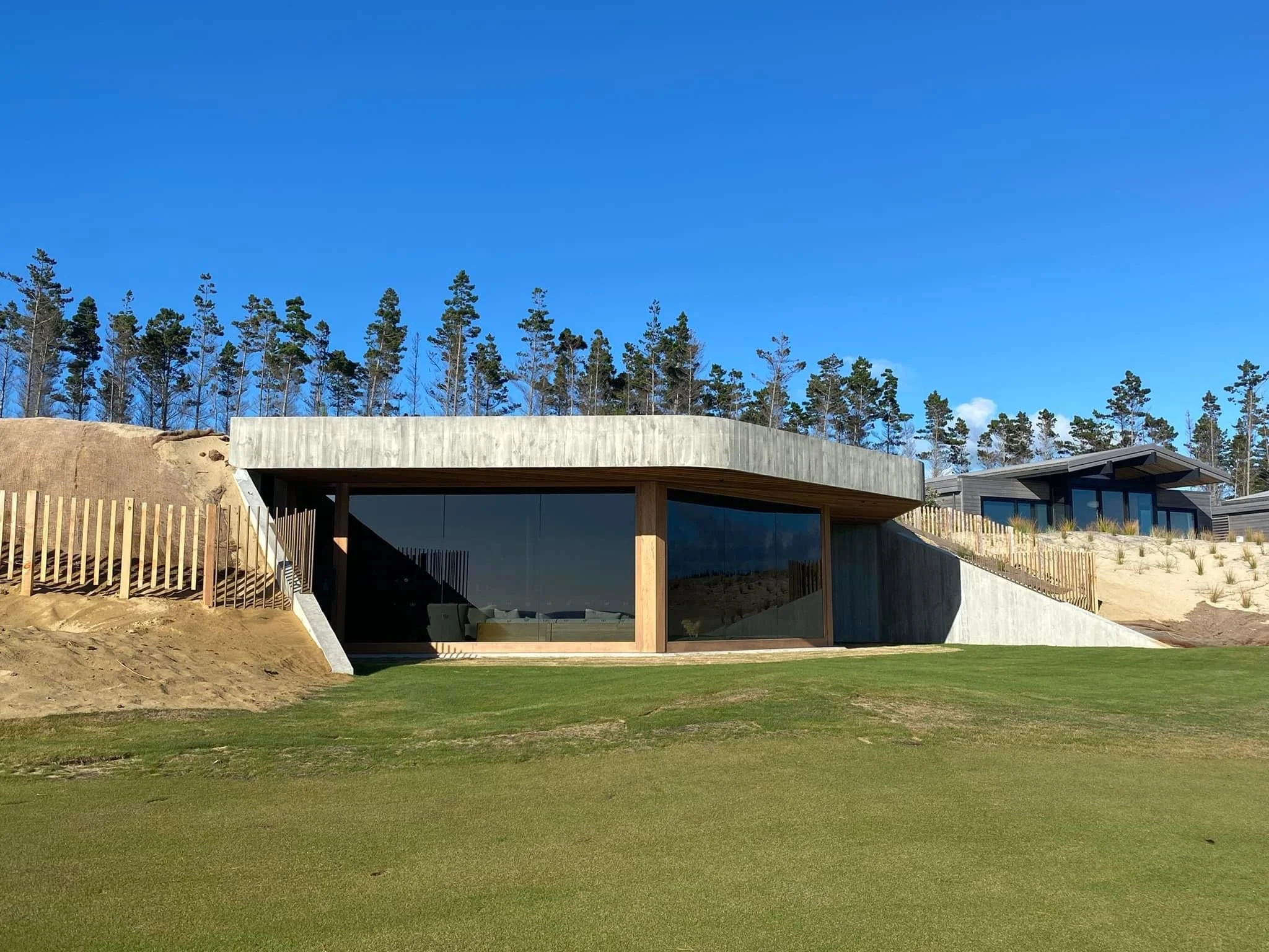 Modern house with large glass windows, concrete and wood exterior, built into a sandy hillside with a grassy lawn in front, surrounded by trees under a clear blue sky.