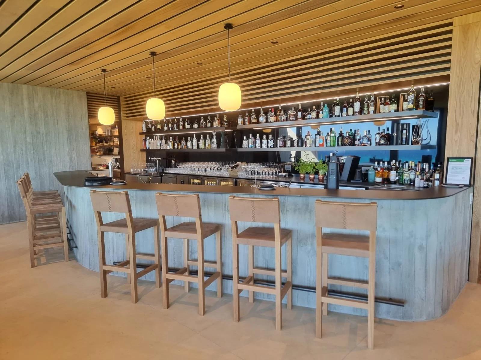 Modern bar with wooden accents, featuring a curved bar counter, four wooden bar stools, illuminated pendant lights, and shelves of liquor bottles behind the counter.