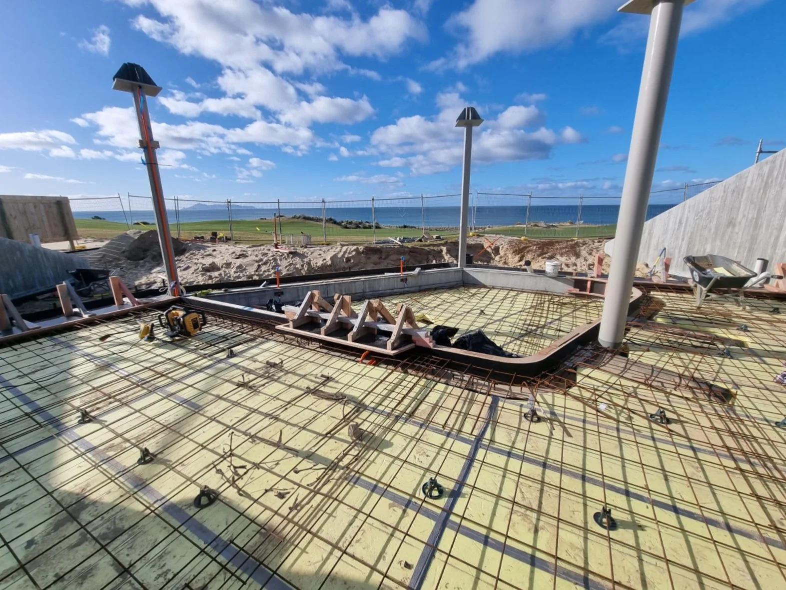 Construction site with rebar and wooden formwork, overlooking a beach and ocean under blue sky with scattered clouds. The Bunker Bar Te Arai Links.