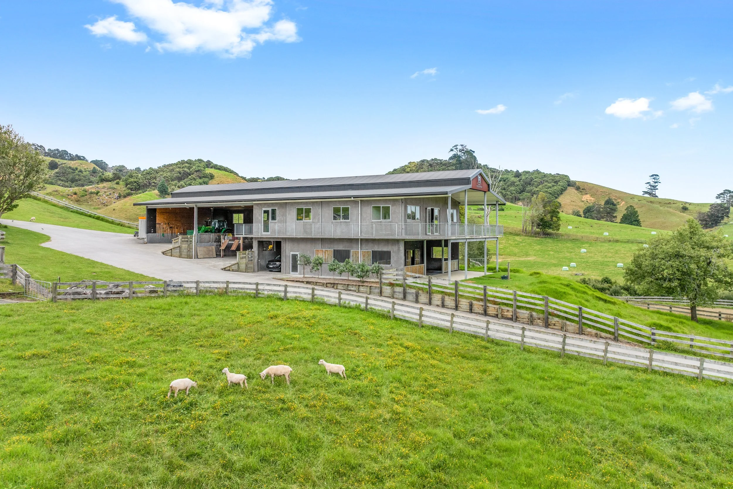 A large, modern barn on a green hillside with grazing sheep in the foreground, under a bright blue sky with a few white clouds.