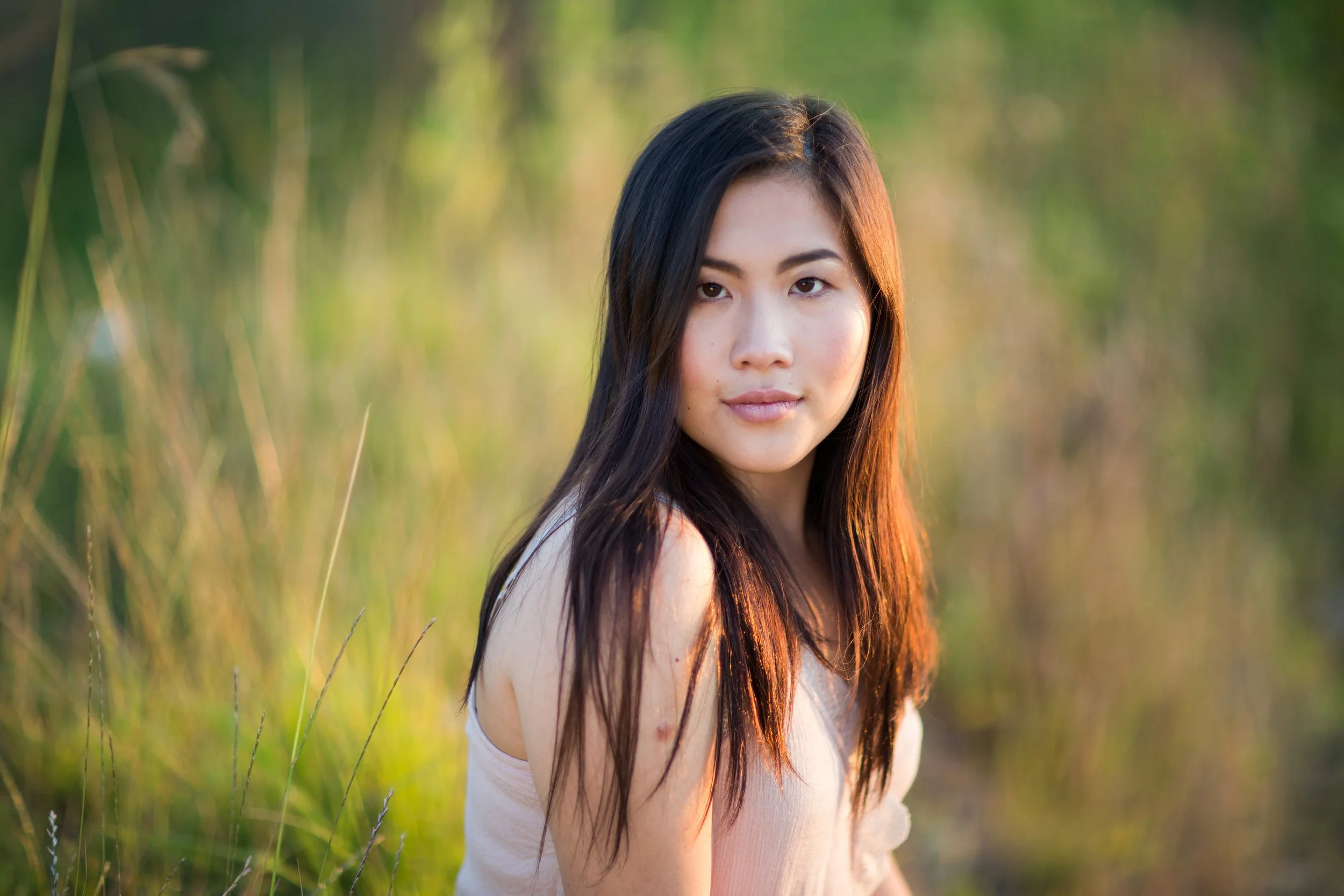 A woman with long dark hair and light skin gazing at the camera in a field of tall grass during sunset.