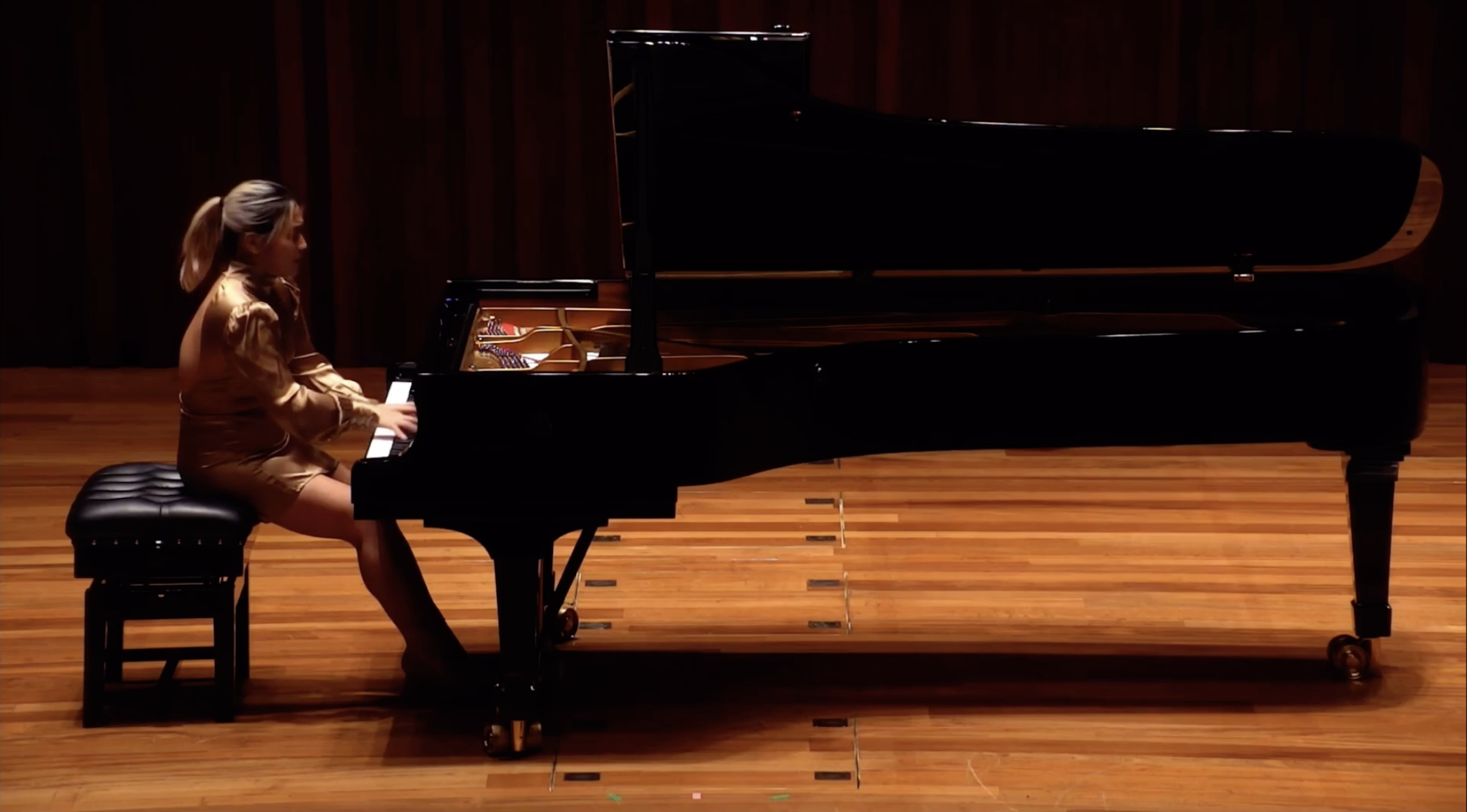 A woman playing a grand piano on a stage with a wooden floor and dark wood paneling background.