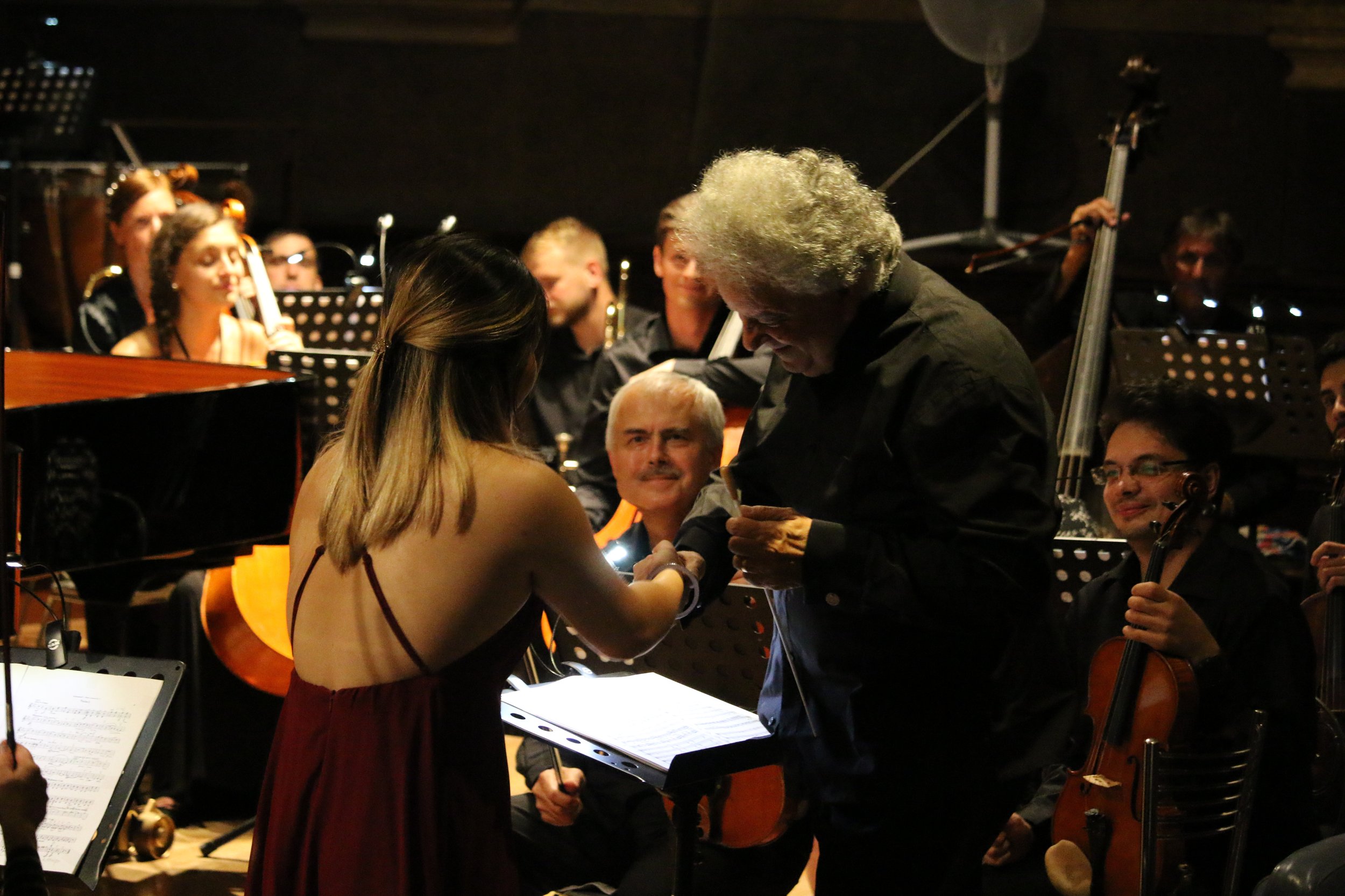 A woman receiving an award or recognition from an older man during a concert or orchestral performance, with an orchestra in the background.