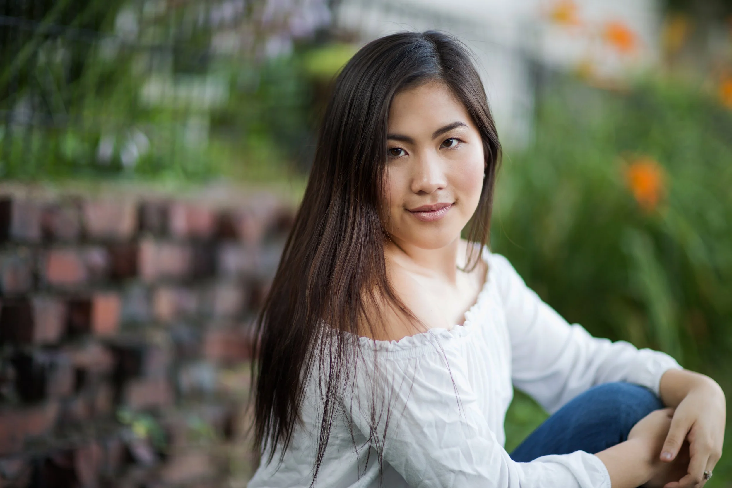 A young woman with long dark hair, wearing a white top, sitting outdoors in a garden with green plants and a brick wall in the background.