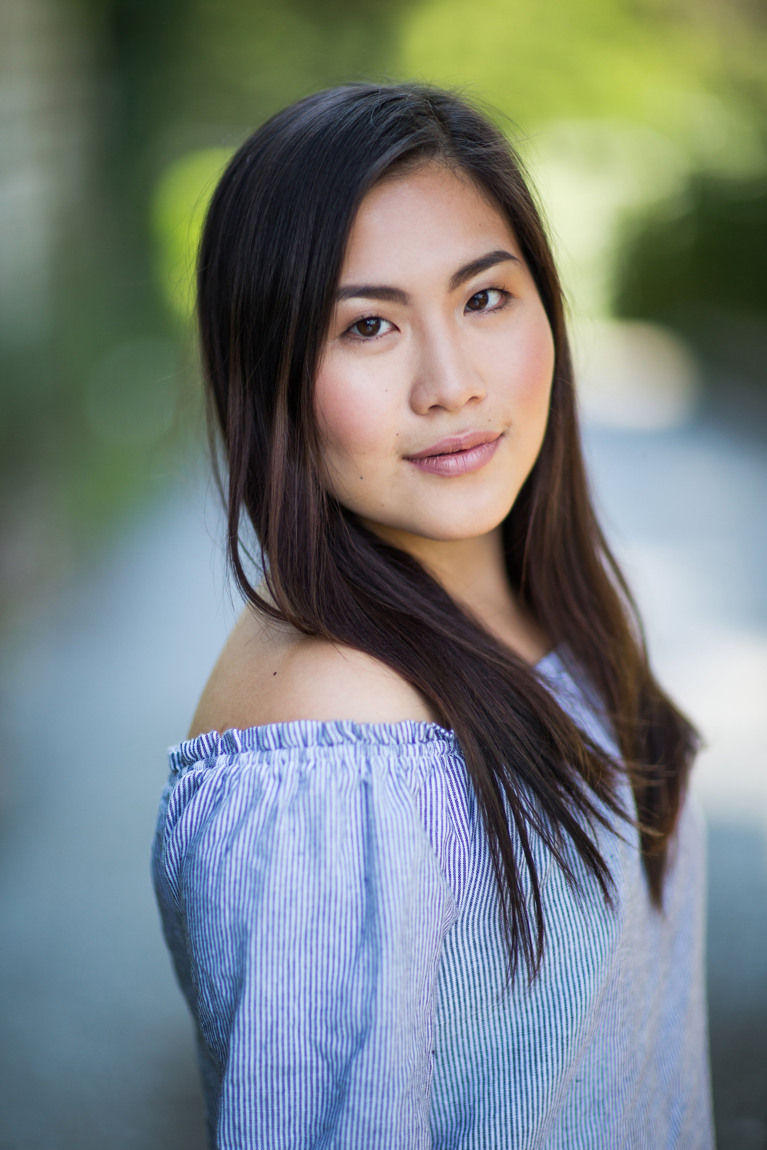 A young woman with long dark hair, wearing a blue and white off-shoulder top, outdoors with a blurred green background.