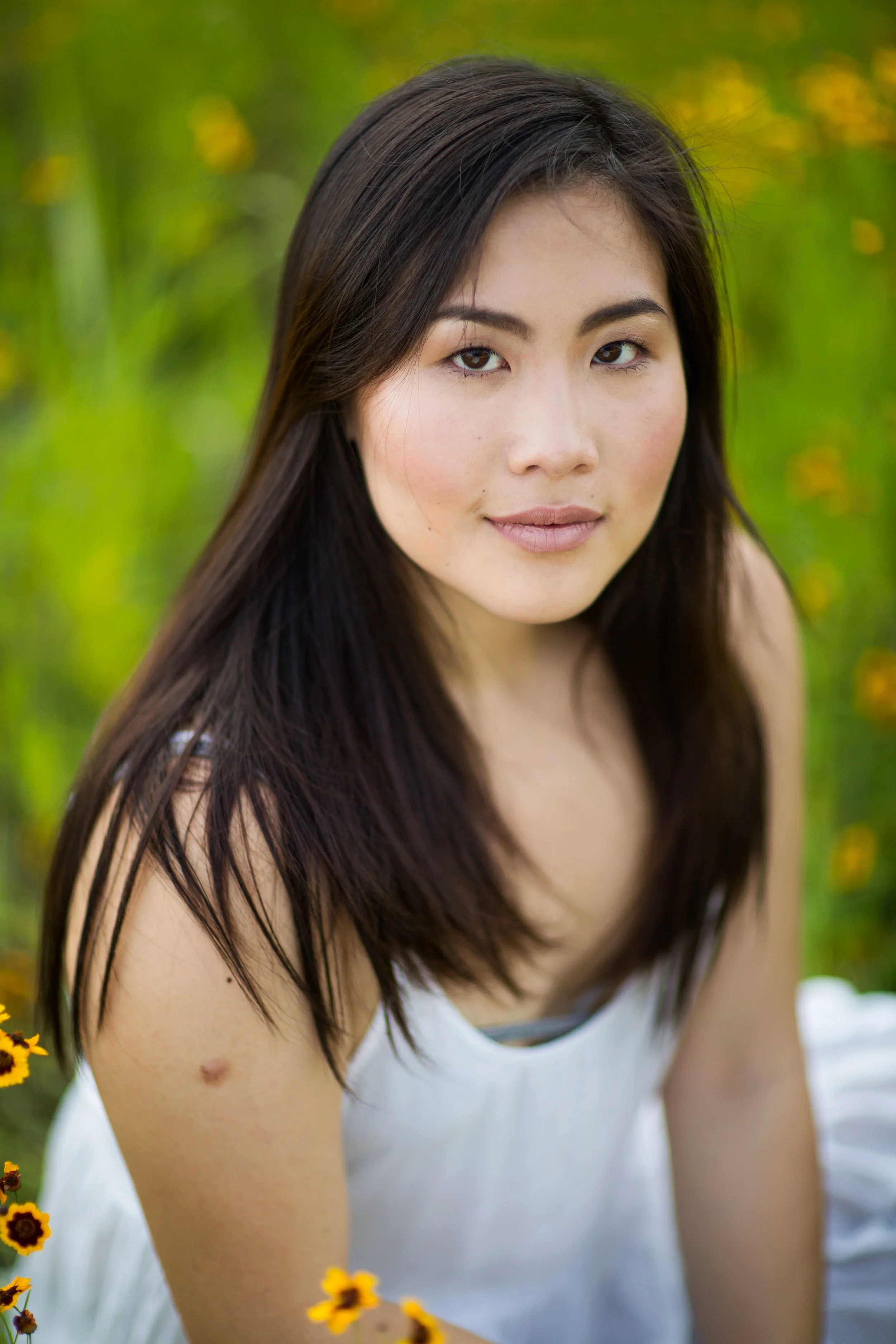 A young woman with long dark hair, wearing a sleeveless white top, sitting outdoors in a field with yellow flowers and green foliage, looking into the camera.