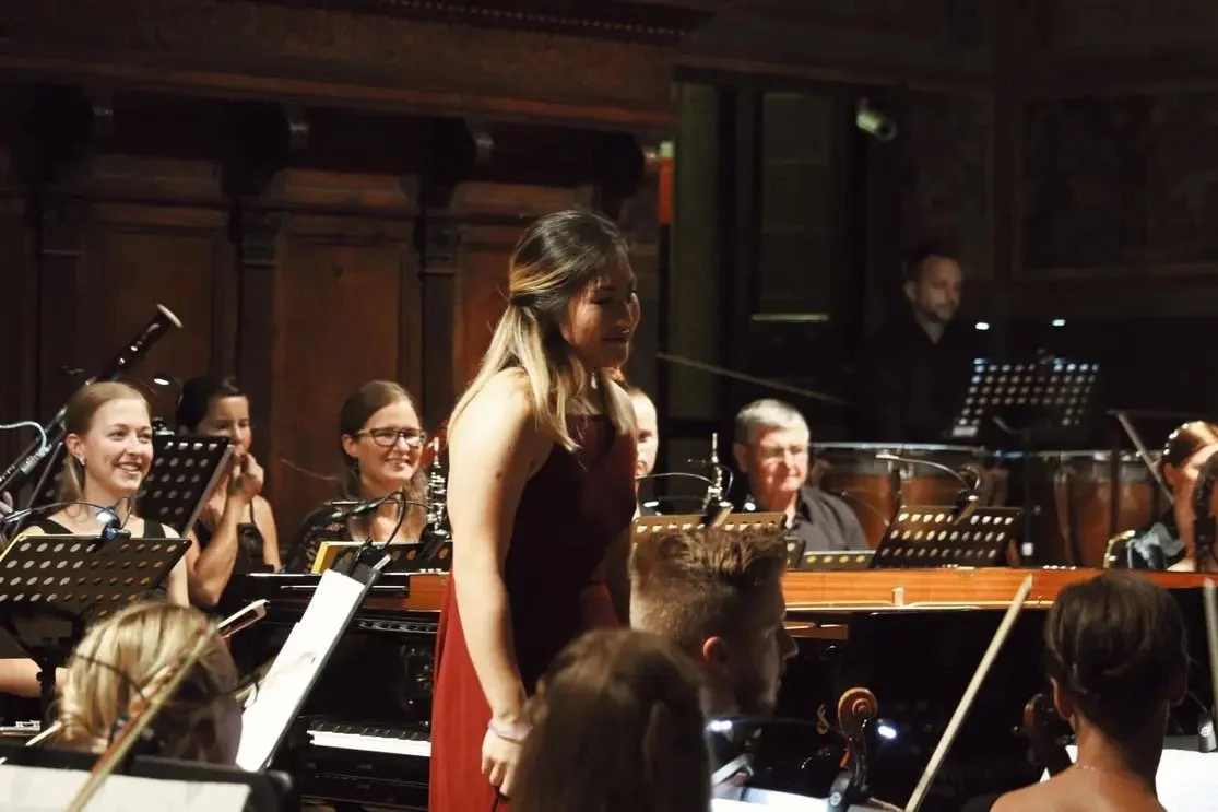 A group of musicians performing in an orchestra, with a woman in a red dress standing in front of them, smiling.