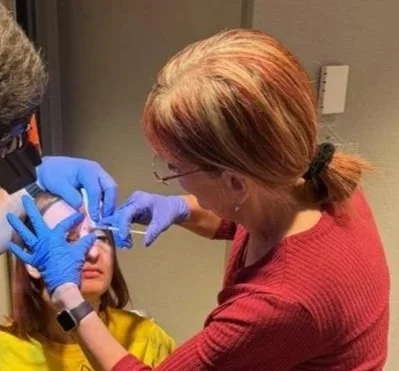 A woman with red hair and glasses administering a dental or medical procedure to a young girl, both wearing gloves.
