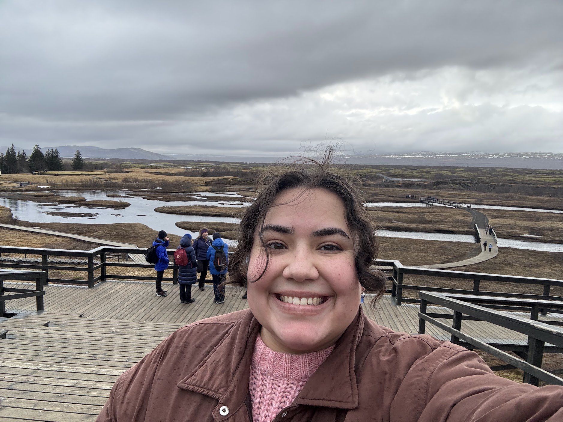 Emma smiling for a selfie with a scenic Icelandiclandscape, winding boardwalk, and cloudy sky in the background.