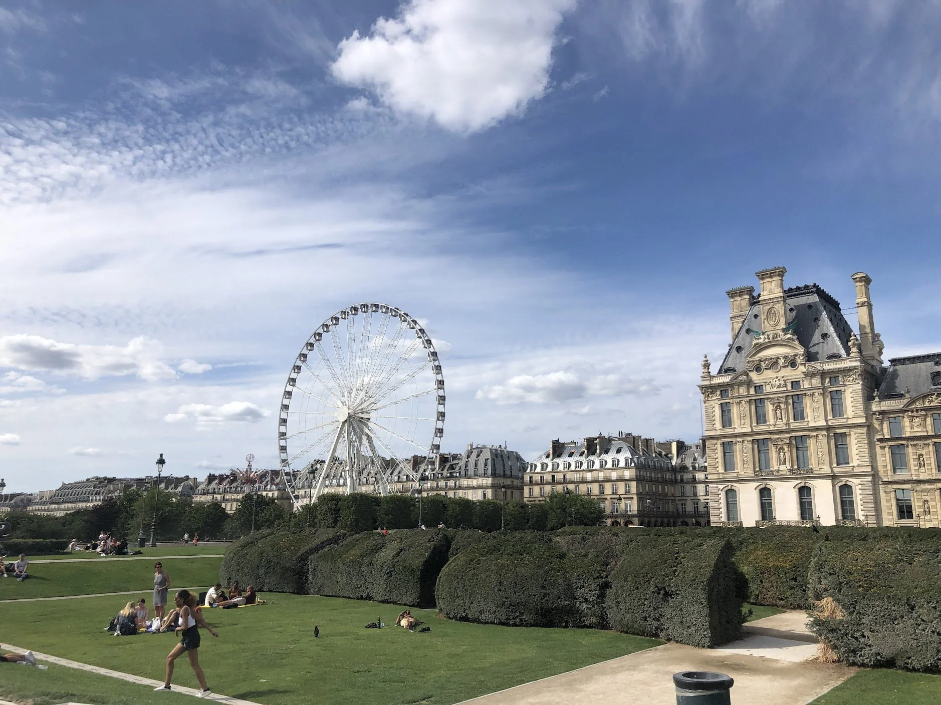 Scene of a park with people sitting and walking on the grass, a large Ferris wheel in the background, and a historic building on the right, under a partly cloudy sky.