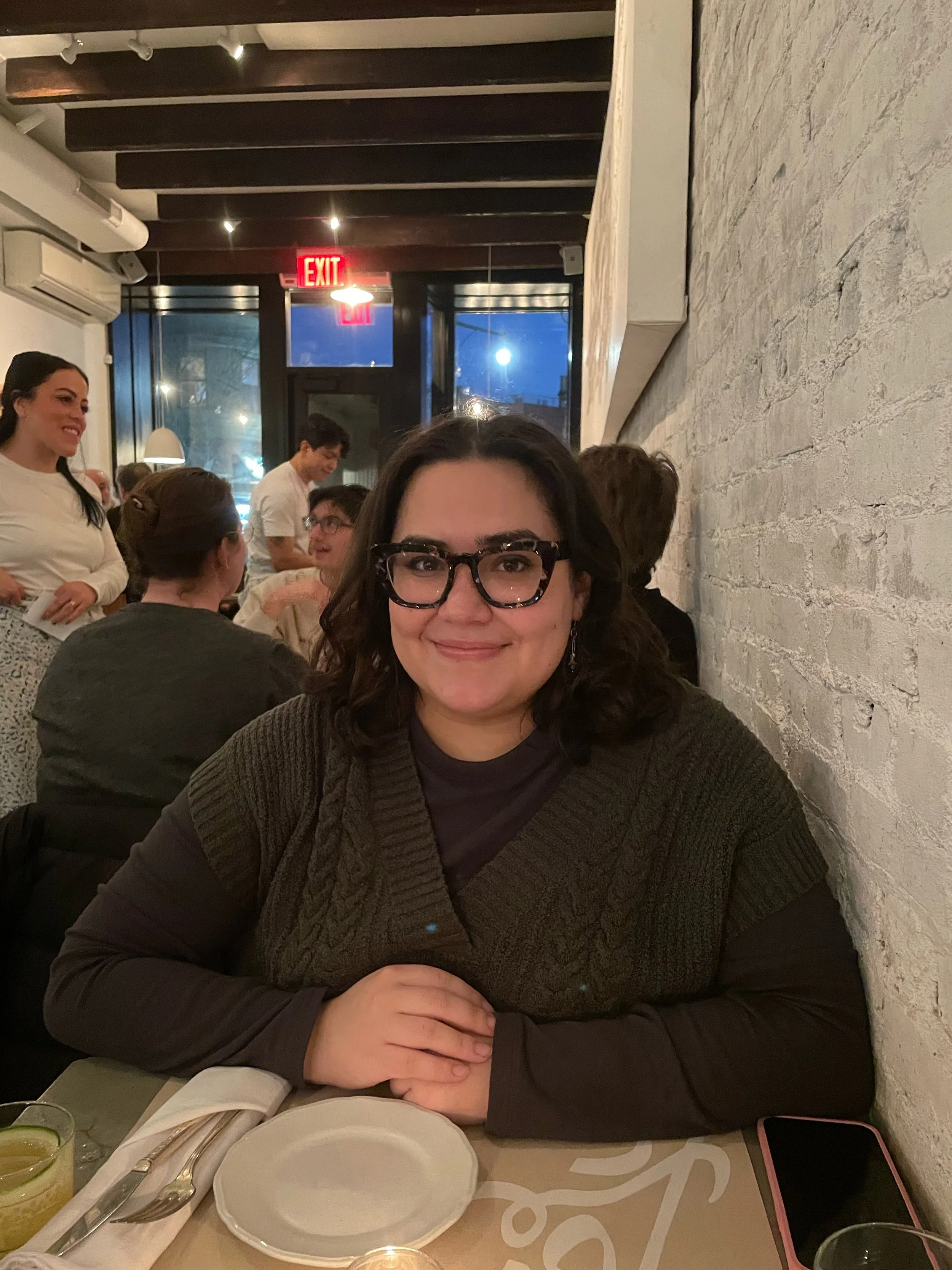 A latina woman with dark hair and glasses sitting at a restaurant table, smiling at the camera. There are other people and a brick wall in the background.