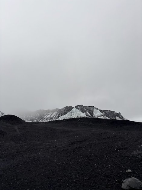 A barren, black volcanic landscape with snow-capped mountains in the distance under a gray, overcast sky.