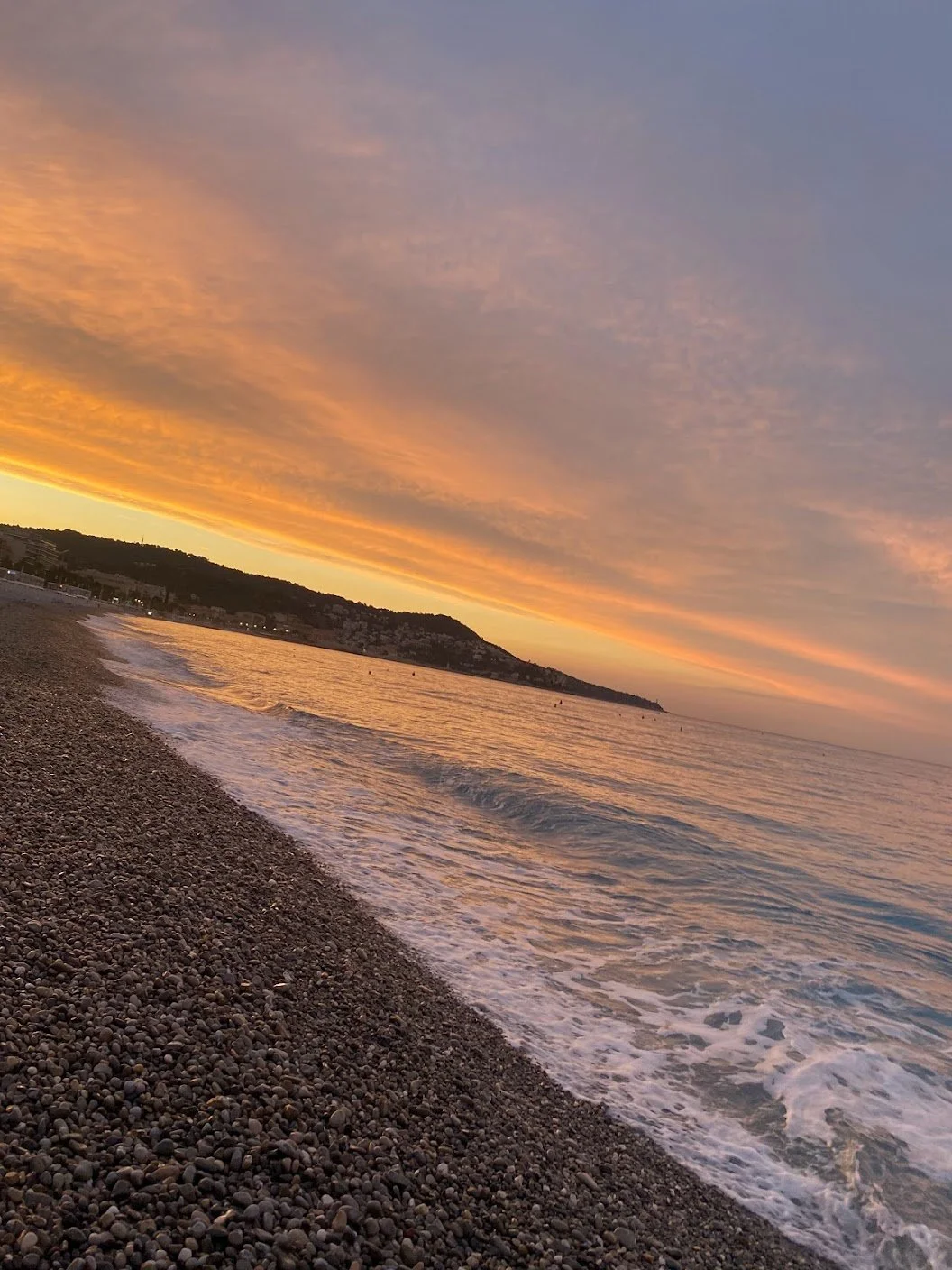 Sunset over a pebble beach with waves gently lapping the shore, colorful clouds in the sky, and a distant view of a hilly area.
