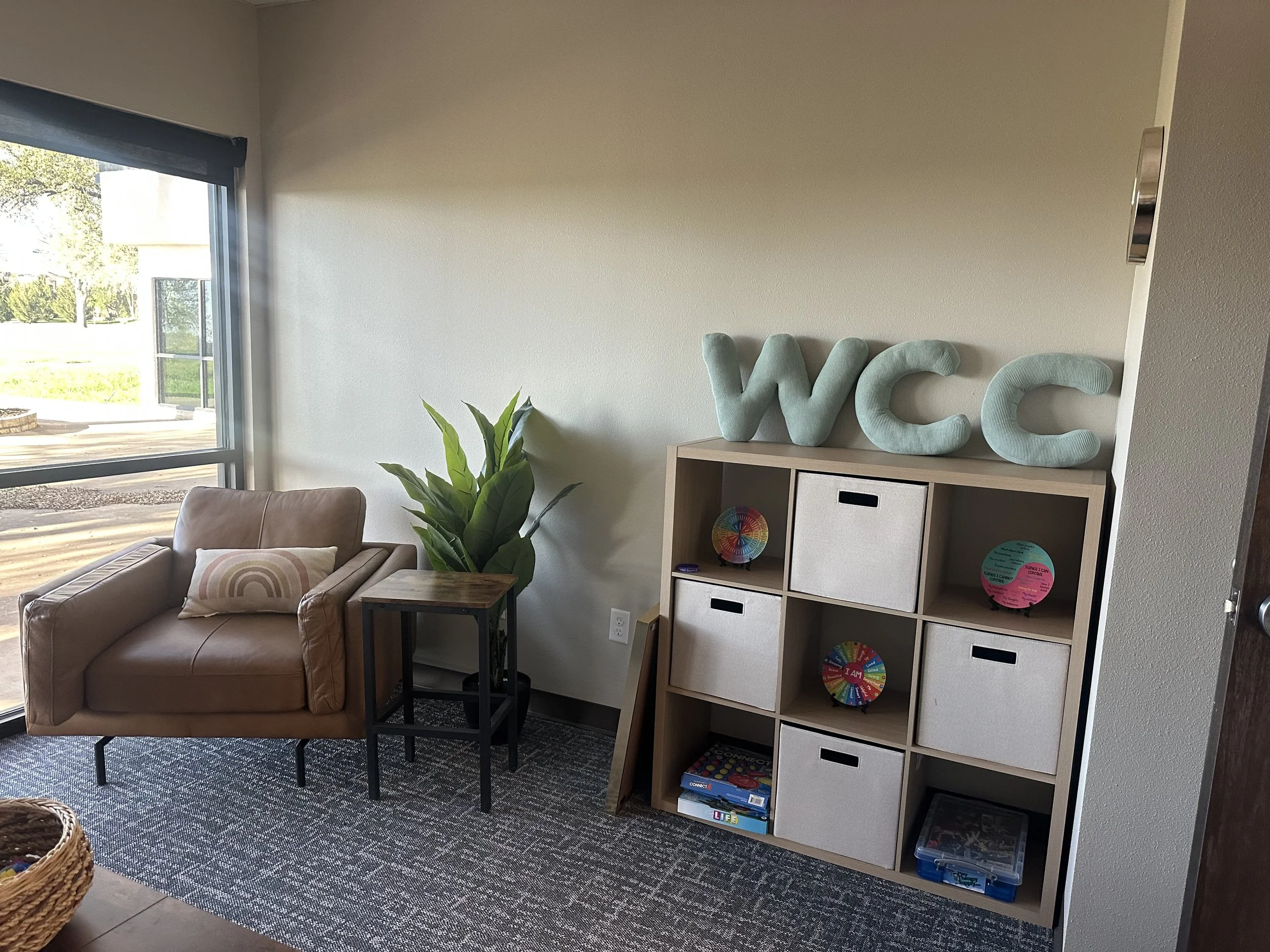 Living room with beige sofa, small side table, indoor plant, white storage cubbies with various items including colorful boards, next to large window showing outdoor green area.