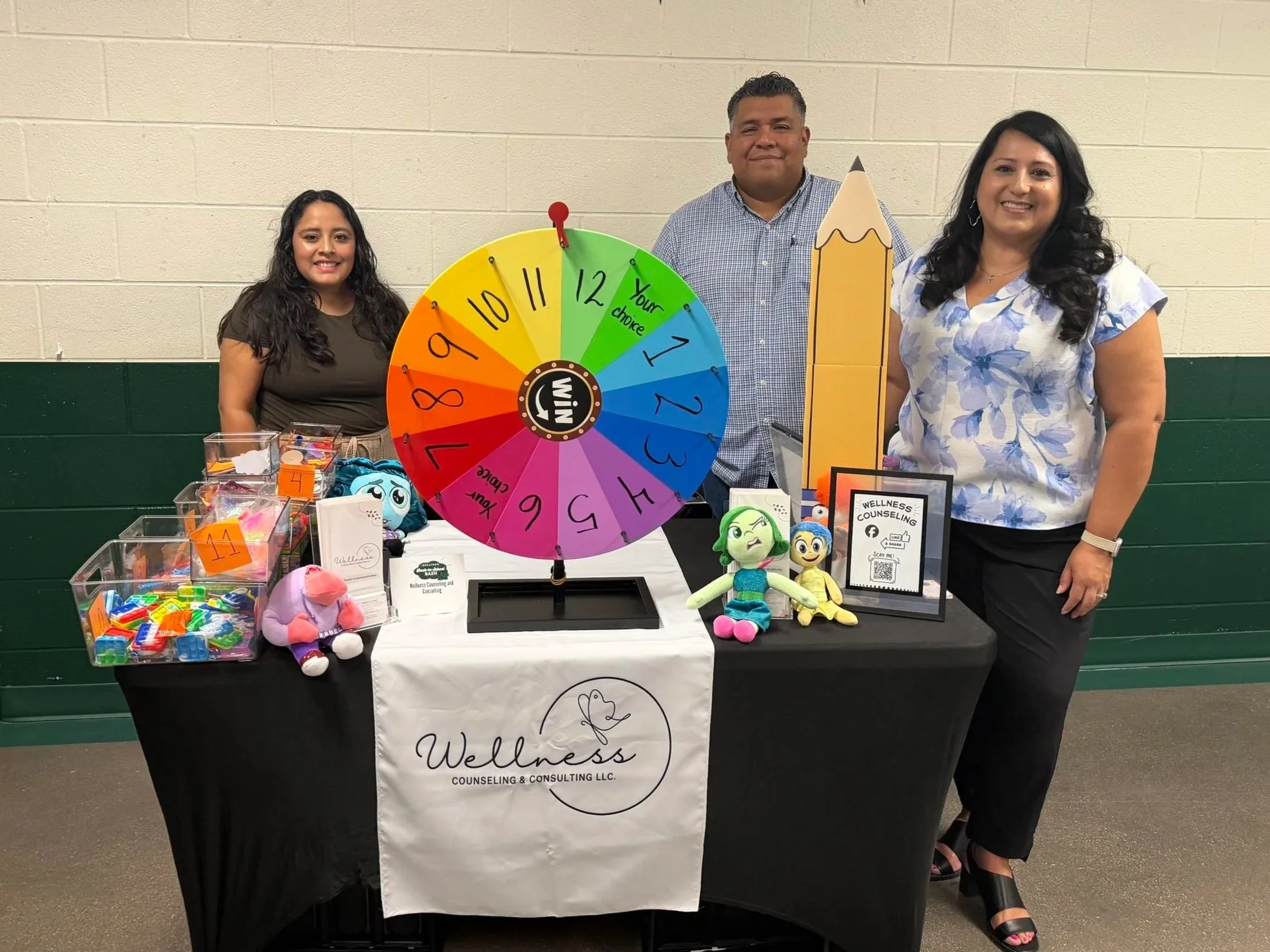 Photo of three staff members and counselors standing at an event, with a large colorful wheel labeled 'Win' and other items including plush toys, small containers, and promotional materials in front of them, against a green and beige wall.
