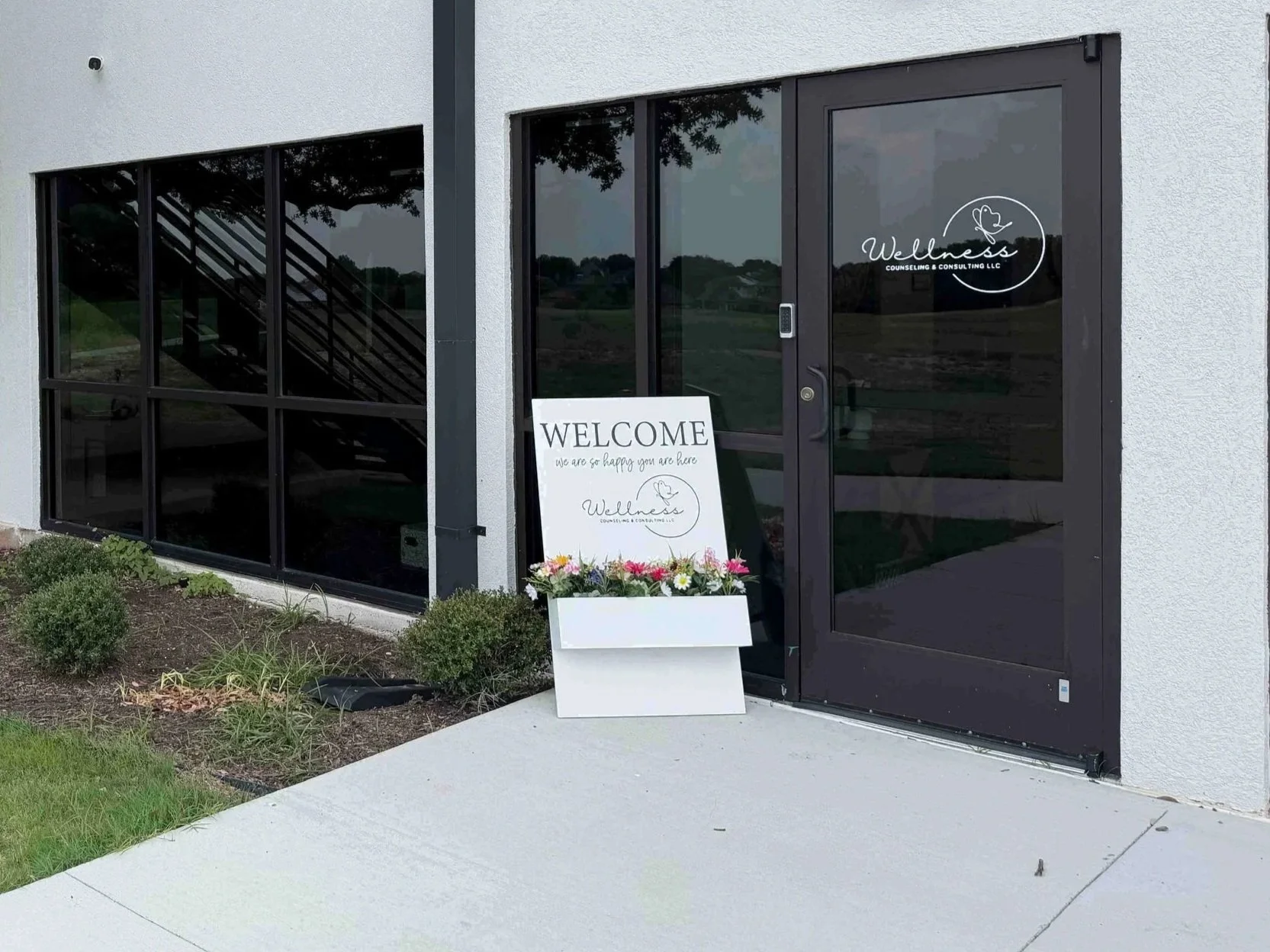 Exterior entrance to a wellness counseling center with glass doors and windows. A white sign with the logo of Wellness Counseling and Consulting, which is located on the lefthand side of the church building it is housed at the Family Life Center.
