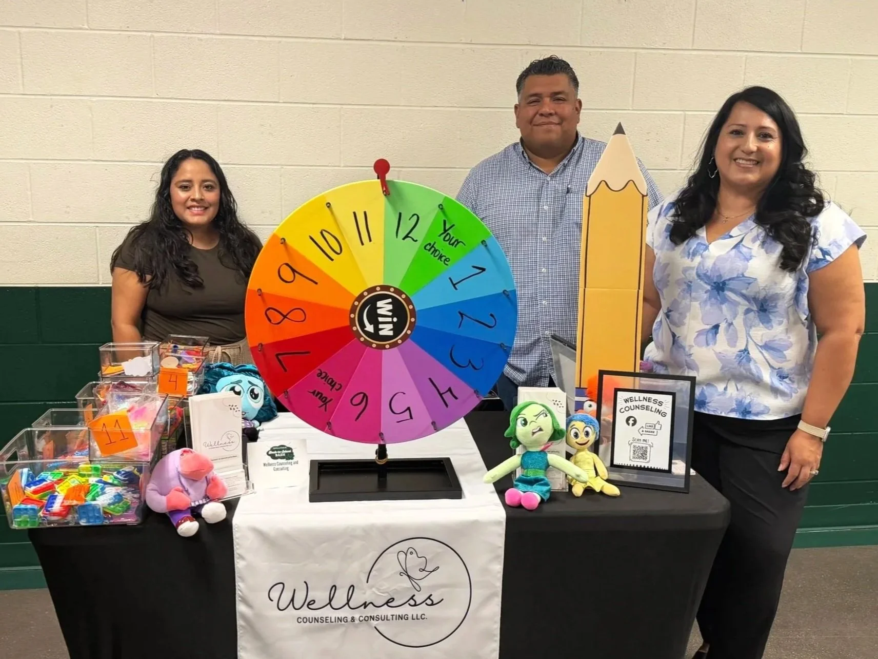 Three counselors standing behind a booth with a colorful prize wheel, toys, and wellness information. The booth displays a banner for Wellness Counseling & Consulting LLC.