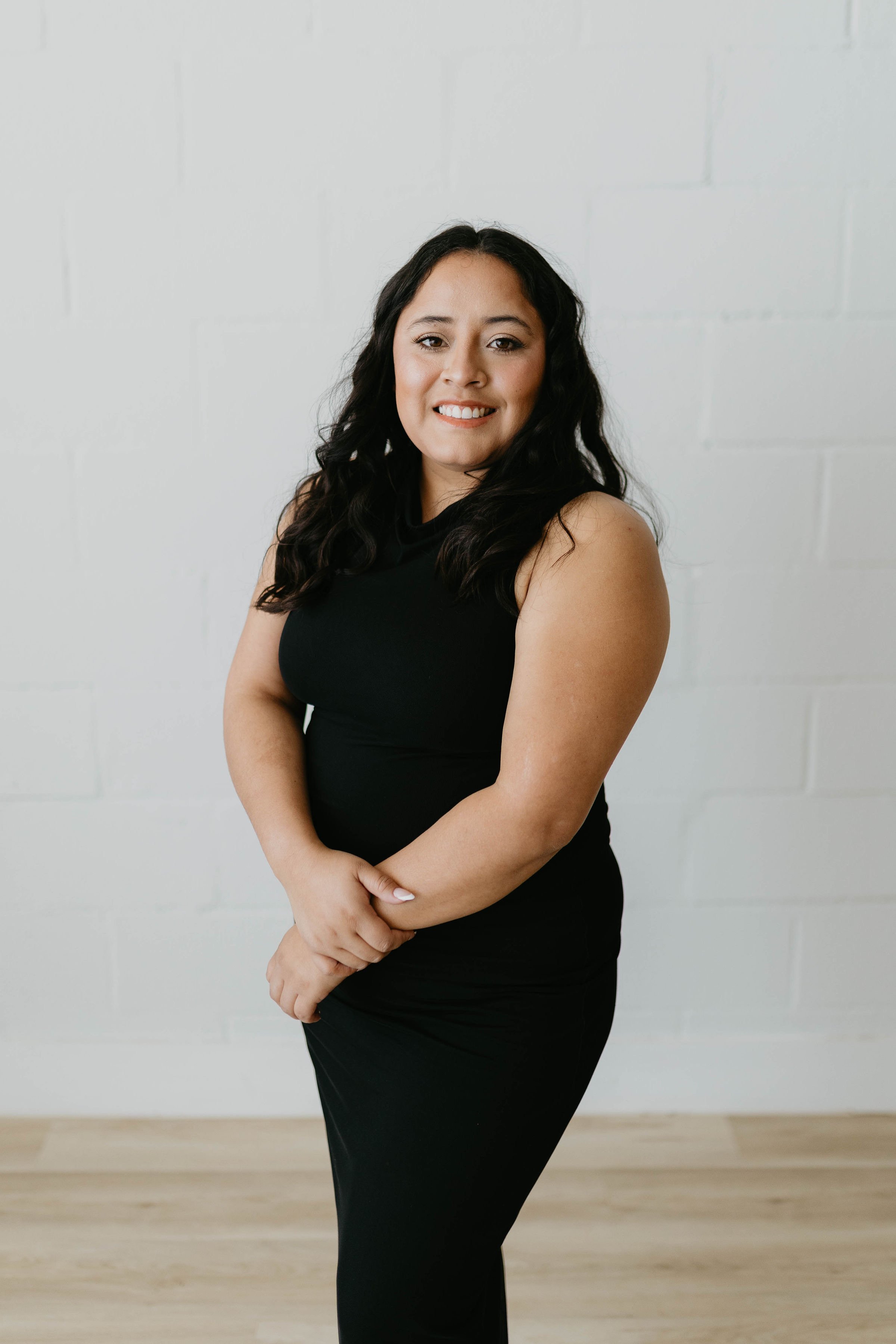 A woman with long dark curly hair wearing a sleeveless black dress standing against a plain white wall, smiling and clasping her hands in front of her.
