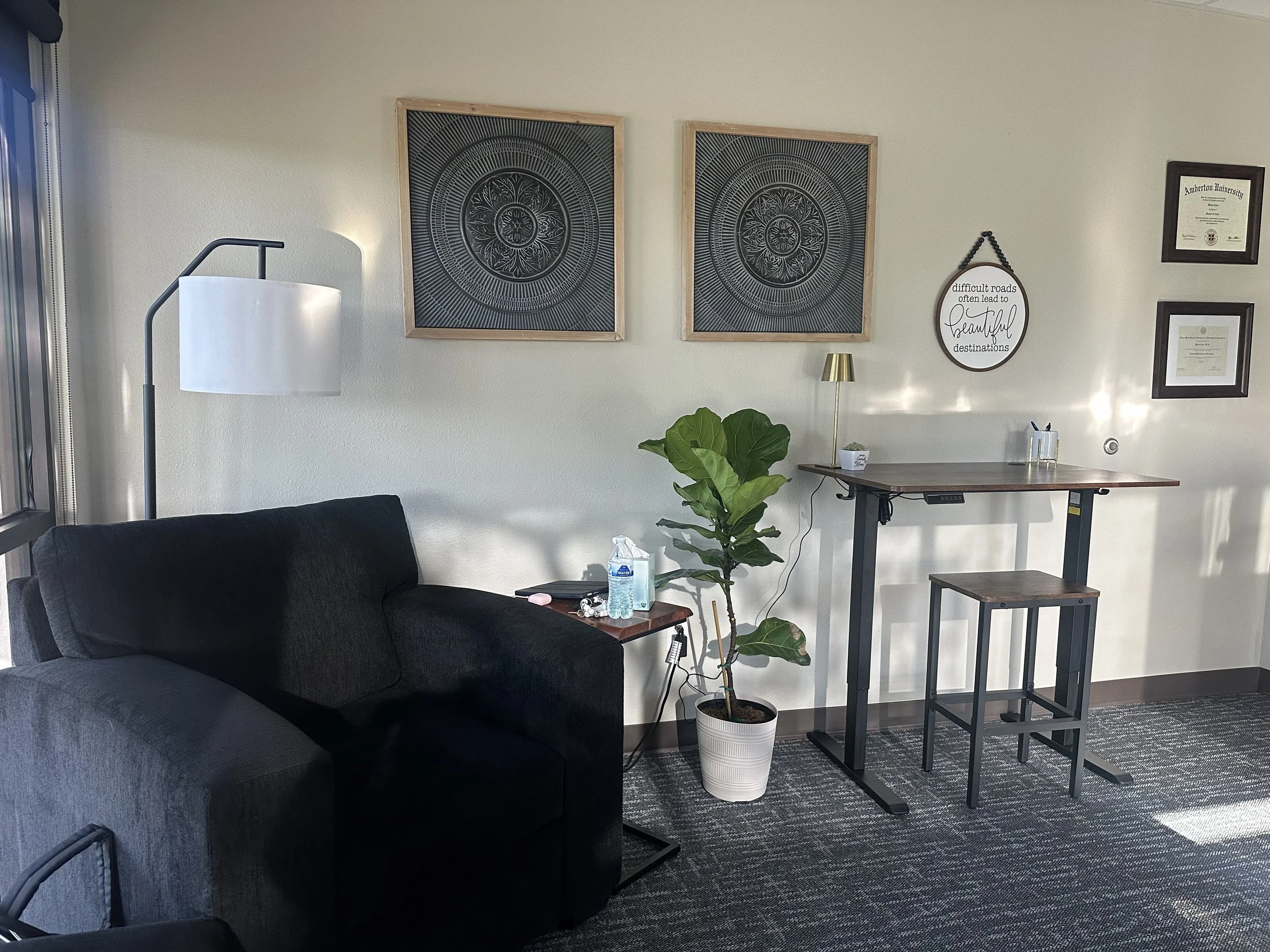 A corner of a room with a black sofa, a potted plant, a small wooden table with a water bottle, a white lamp, and wall art including framed certificates, decorative circular art, and a sign with a quote about roads and destinations.