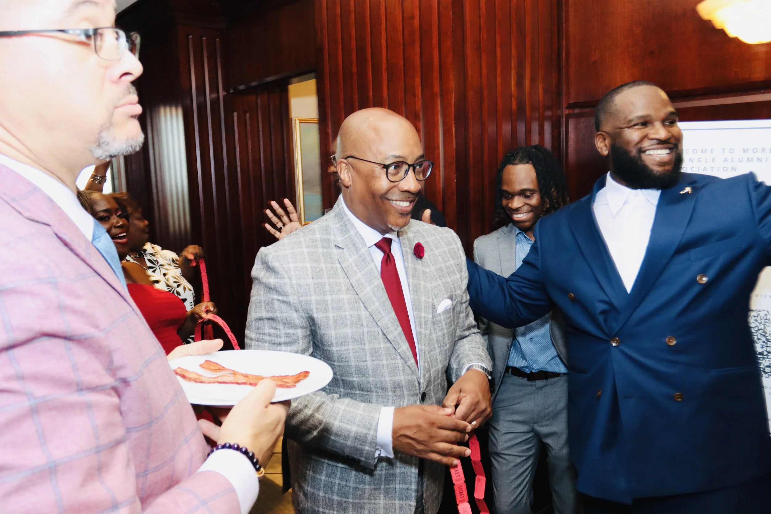 Group of people at a social event, smiling and interacting with each other, with wooden wall paneling in the background.