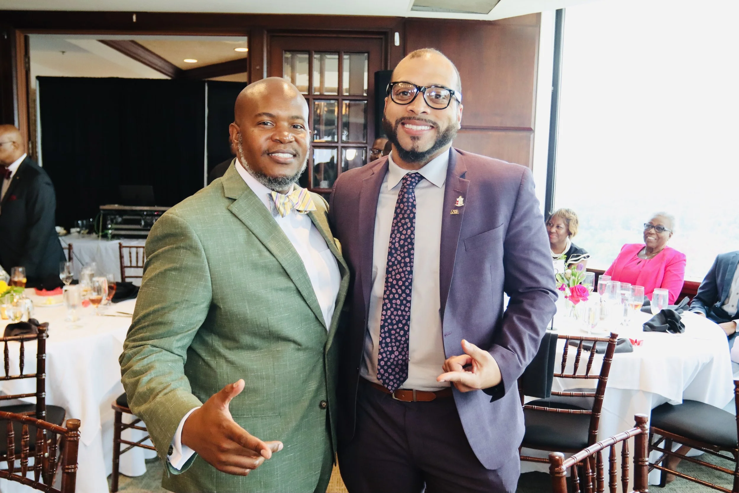 Two men in formal suits smiling and posing for a photo at a banquet or formal event, with women sitting at a table in the background.