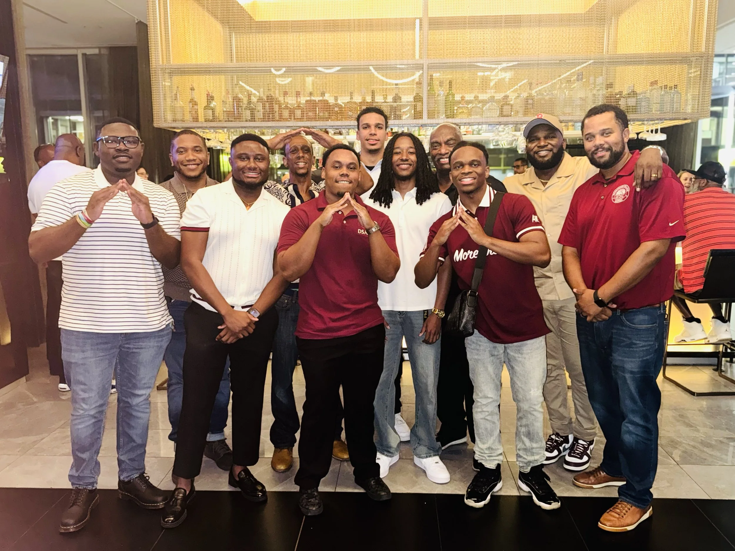 A group of fourteen men standing together inside a restaurant, smiling, some making hand gestures, with a bar area and other patrons in the background.