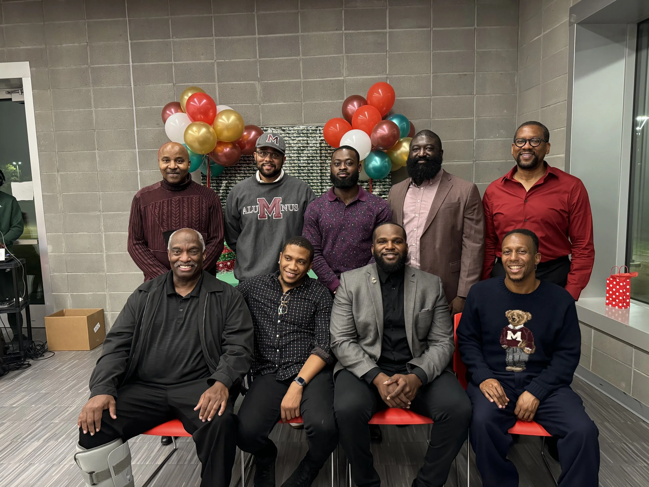 Group of ten men gathered indoors, some sitting and some standing, in front of a wall decorated with balloons and holiday decorations.