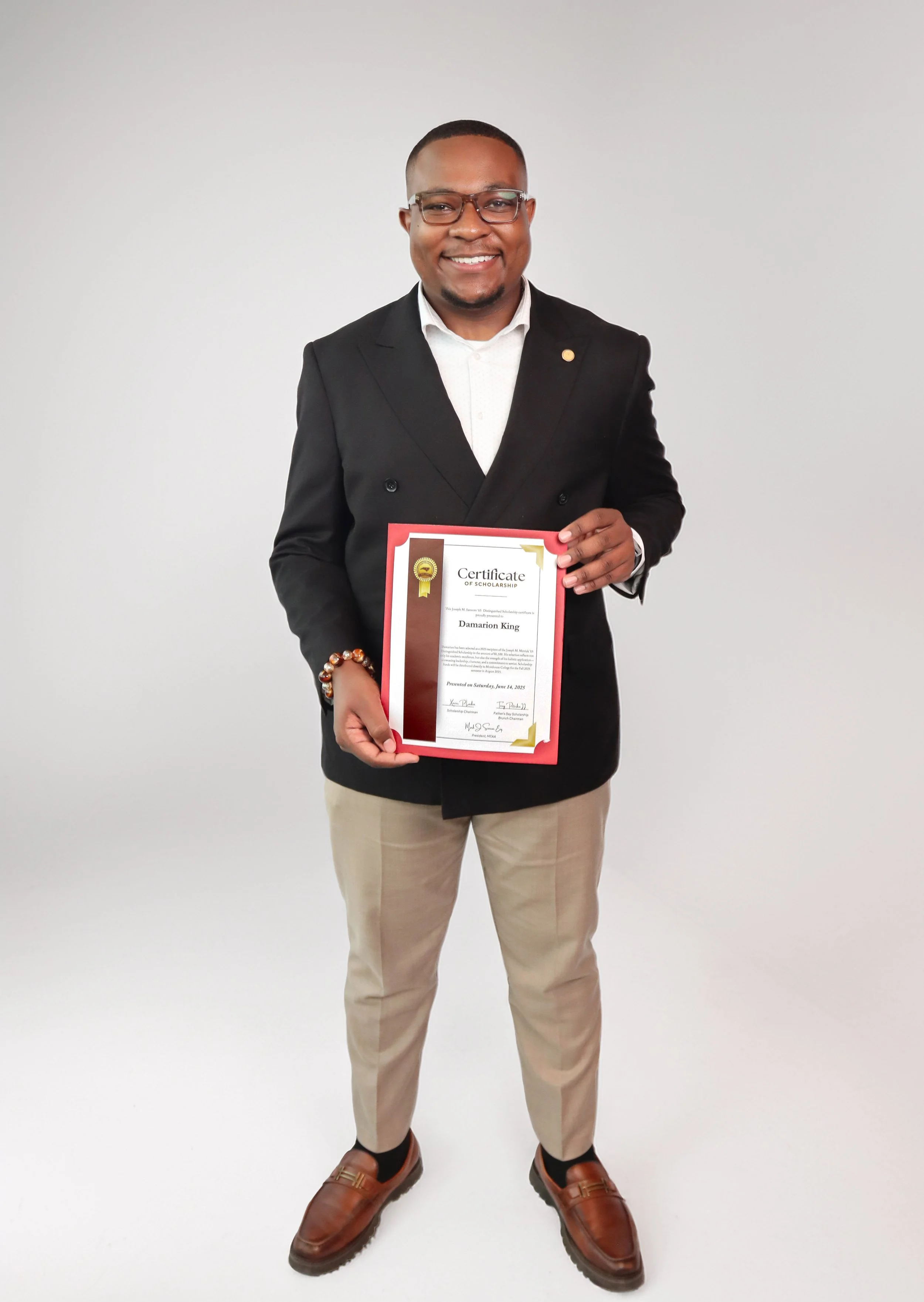 A man in a black blazer, white shirt, and beige pants holding a certificate of scholarship, standing against a plain white backdrop, smiling at the camera.