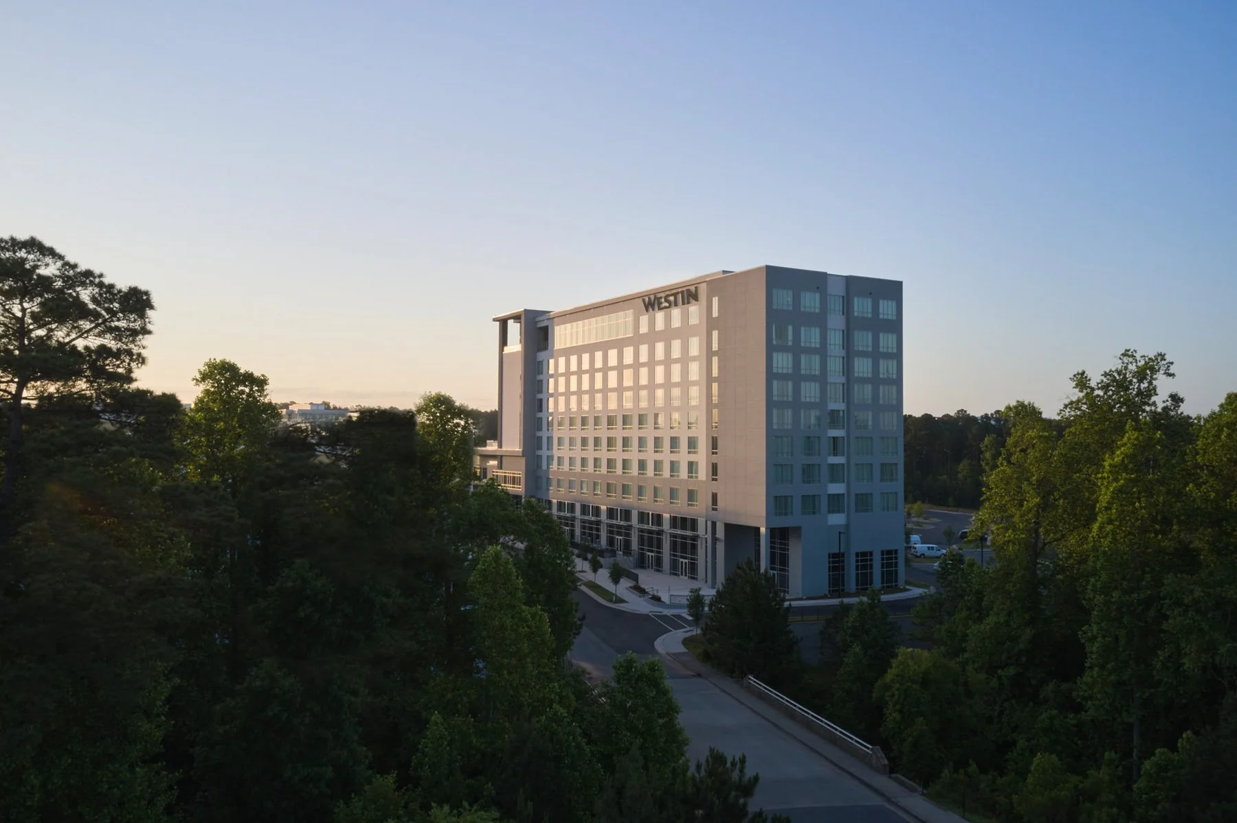 A modern hotel building labeled Westin, surrounded by trees and a parking lot, captured during sunset.