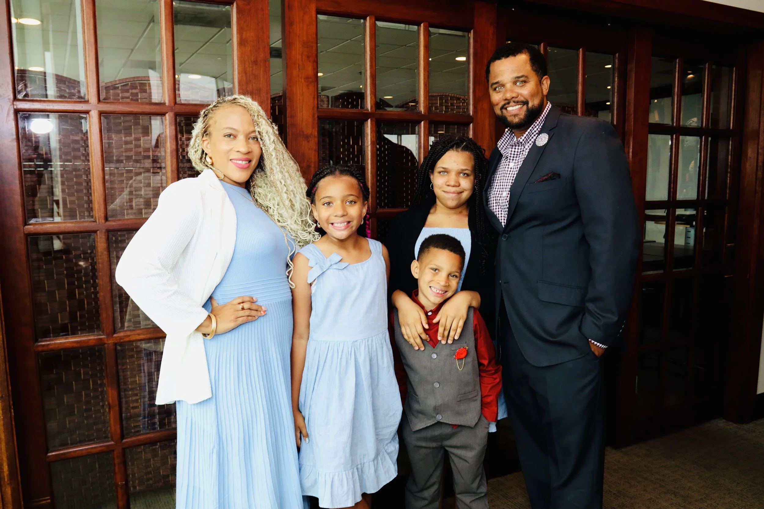 A family of six posing in front of a wooden partition, smiling for the camera at an indoor event.