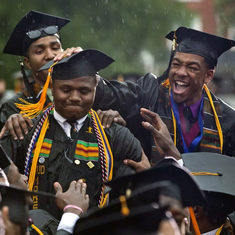 Graduates in caps and gowns celebrating at graduation ceremony, with one person smiling broadly and others in a joyful mood, some holding their caps.