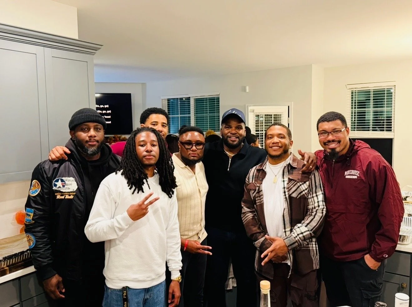 Group of seven men posing together in a kitchen, smiling at the camera, with windows and cabinets in the background.