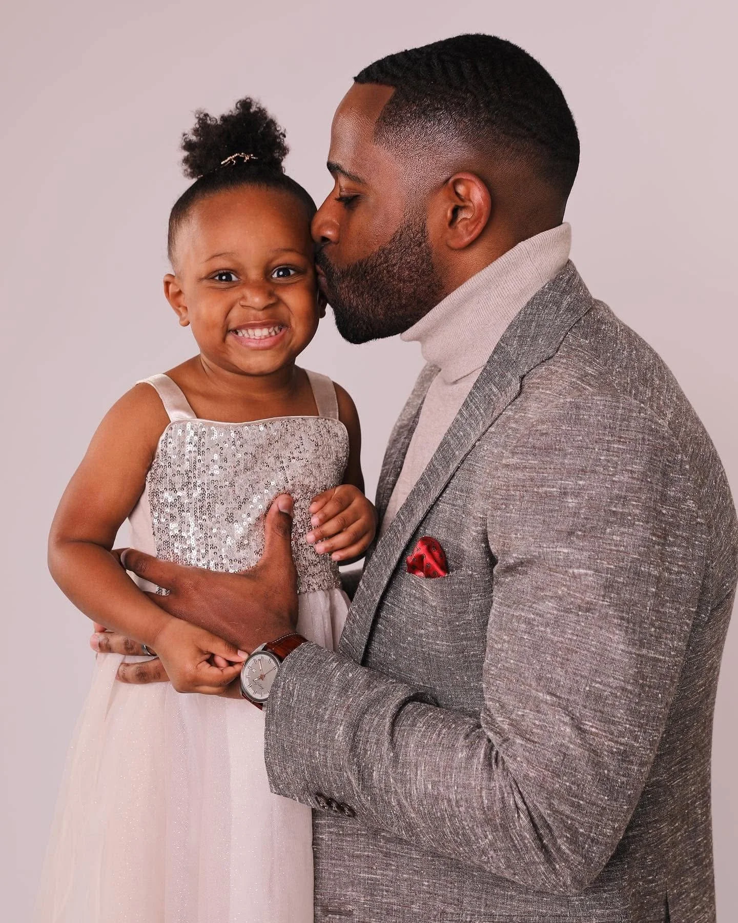 A man in a gray blazer and turtleneck gently holding a young girl, who is smiling and wearing a sparkly dress, as he kisses her forehead.