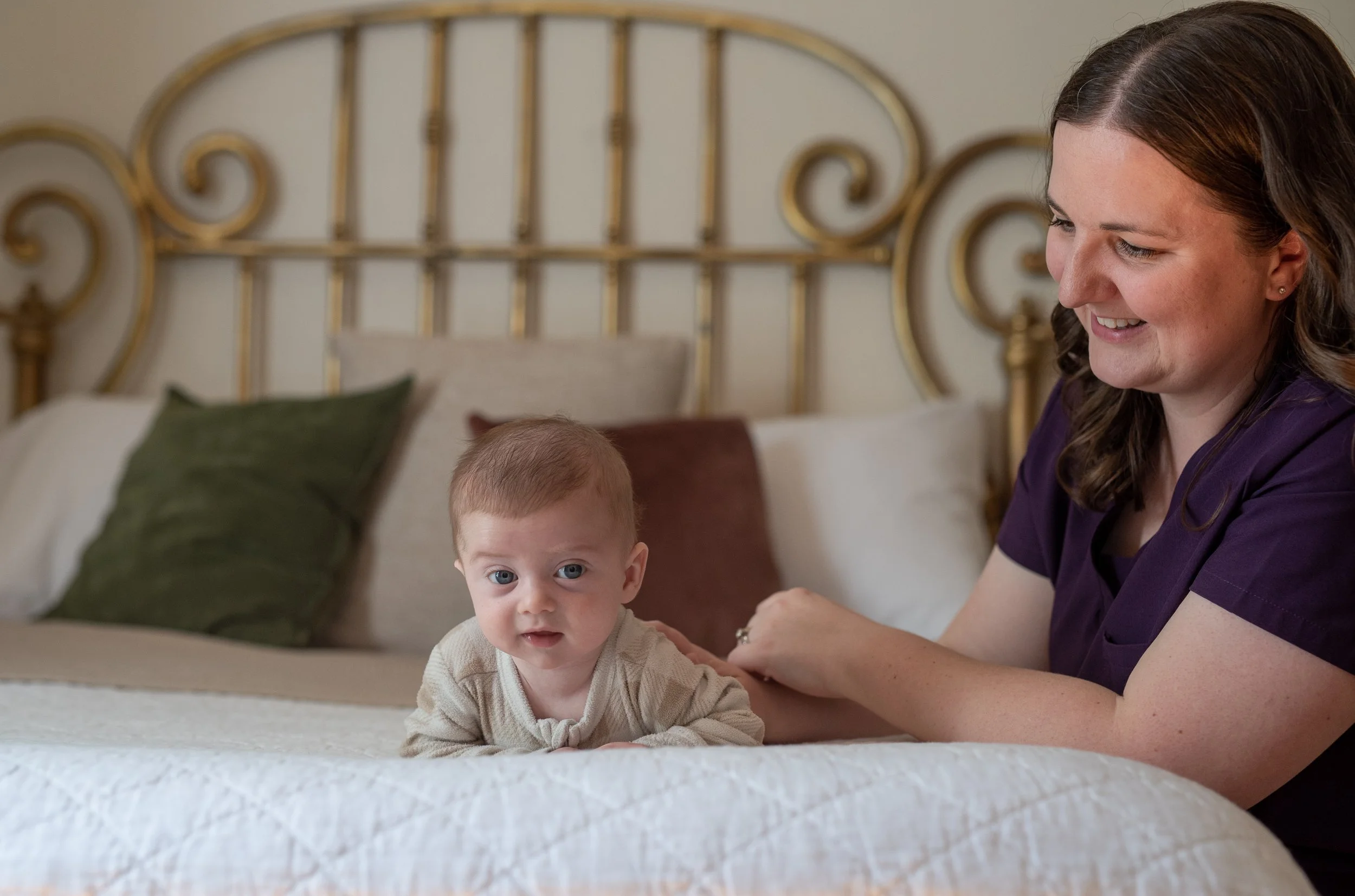 A woman with long brown hair and wearing purple scrubs smiling while looking at a young child with blue eyes and short light-colored hair, lying on a bed with a white textured blanket and green, beige, and brown pillows in the background.