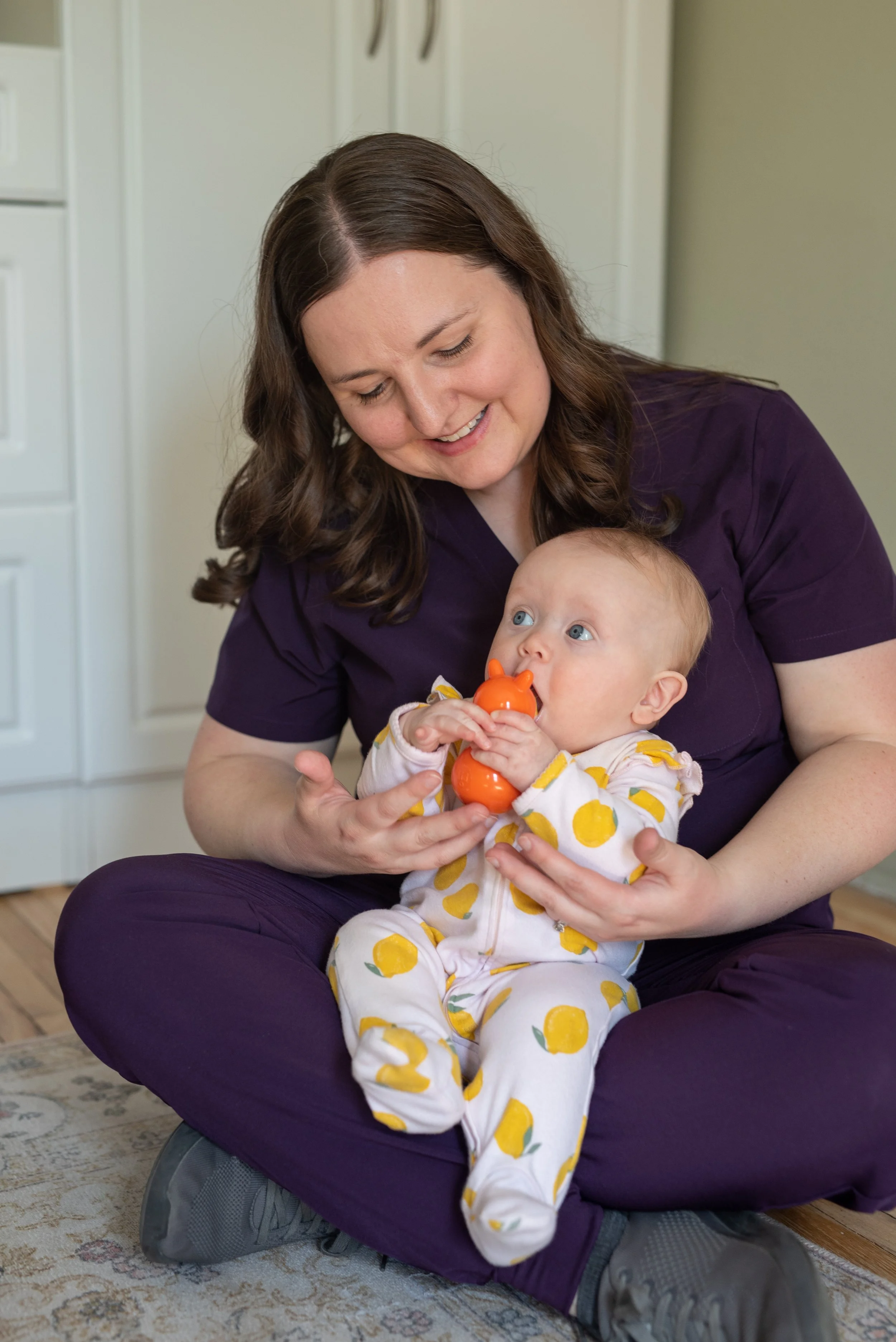 A woman with brown hair, wearing a purple t-shirt and purple pants, is sitting on her knees on a patterned carpet, holding a baby dressed in a lemon-patterned onesie. The woman is smiling and looking at the baby, who is holding an orange sippy cup and looking up with wide blue eyes.