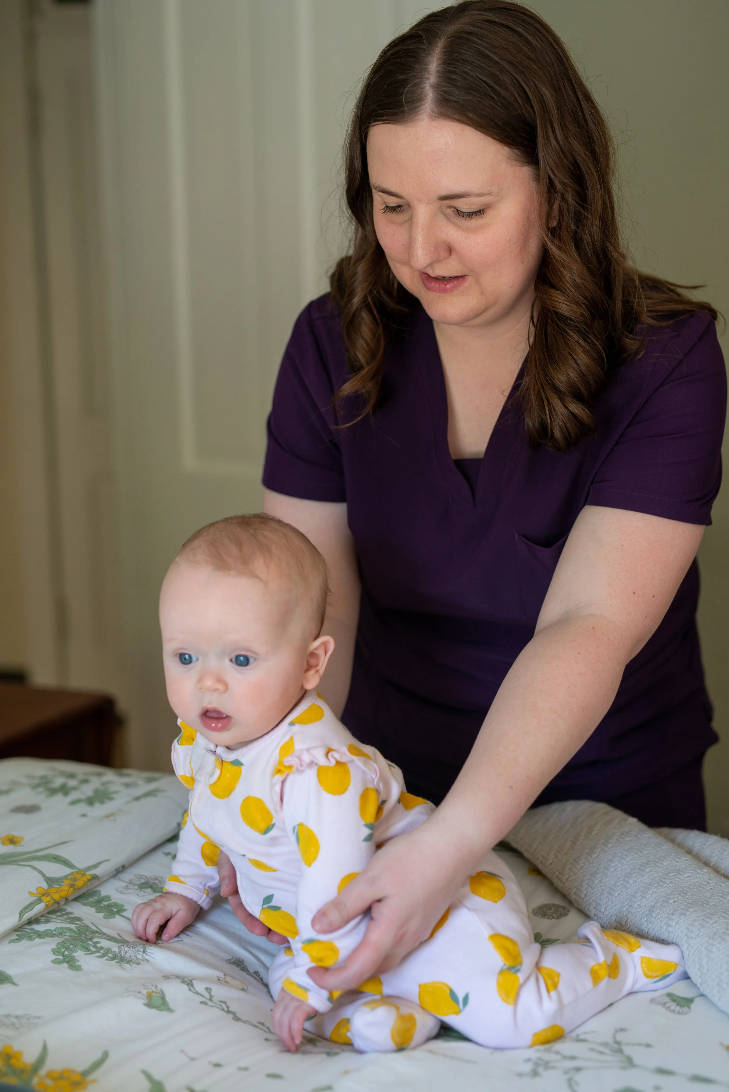 A woman with brown hair and a purple shirt helping a baby with blond hair, wearing a white onesie with yellow lemons, on a bed with a floral bedsheet.