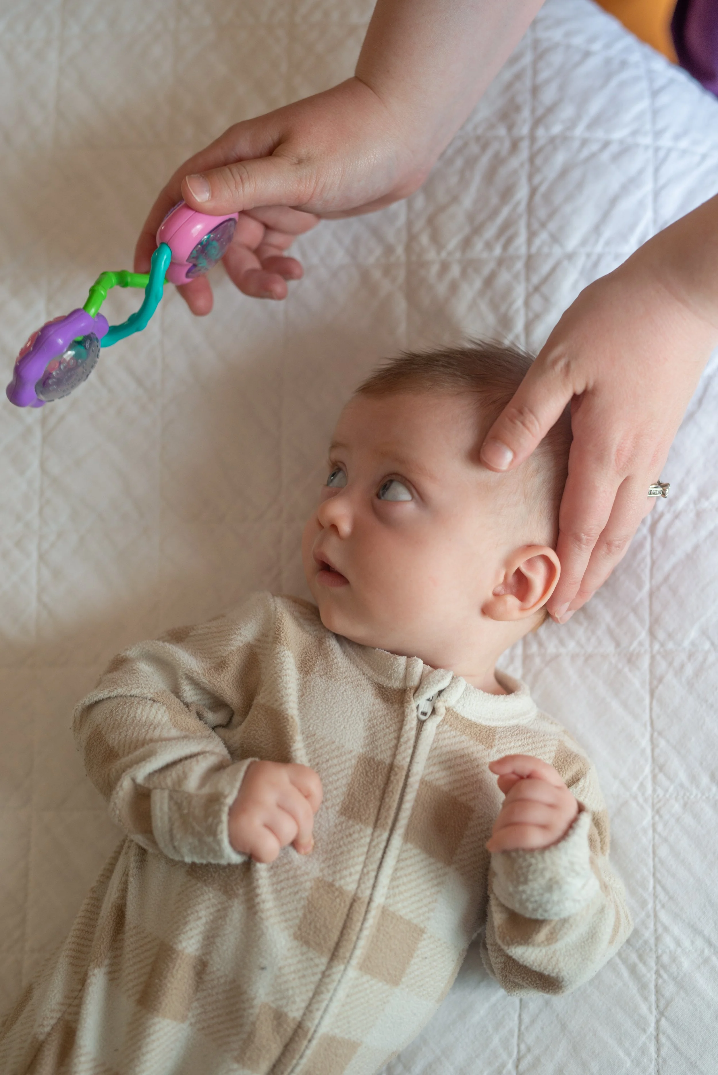 A baby lying on a quilted surface, looking at a toy held by an adult hand. The baby is wearing a beige, zippered hoodie and has their fists clenched.