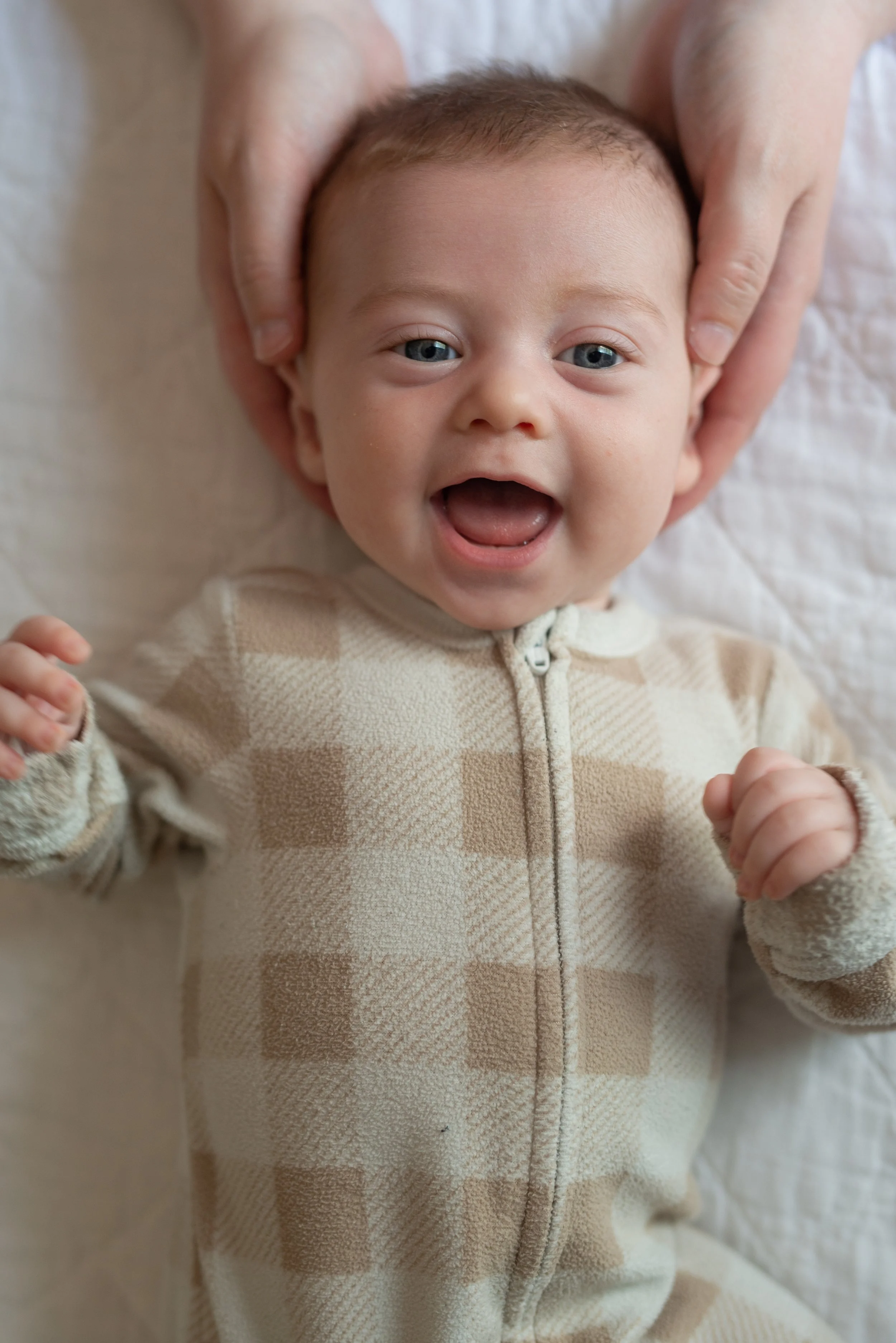 Close-up of a baby with blue eyes smiling and holding fists up, wearing a beige and brown checkered hoodie.