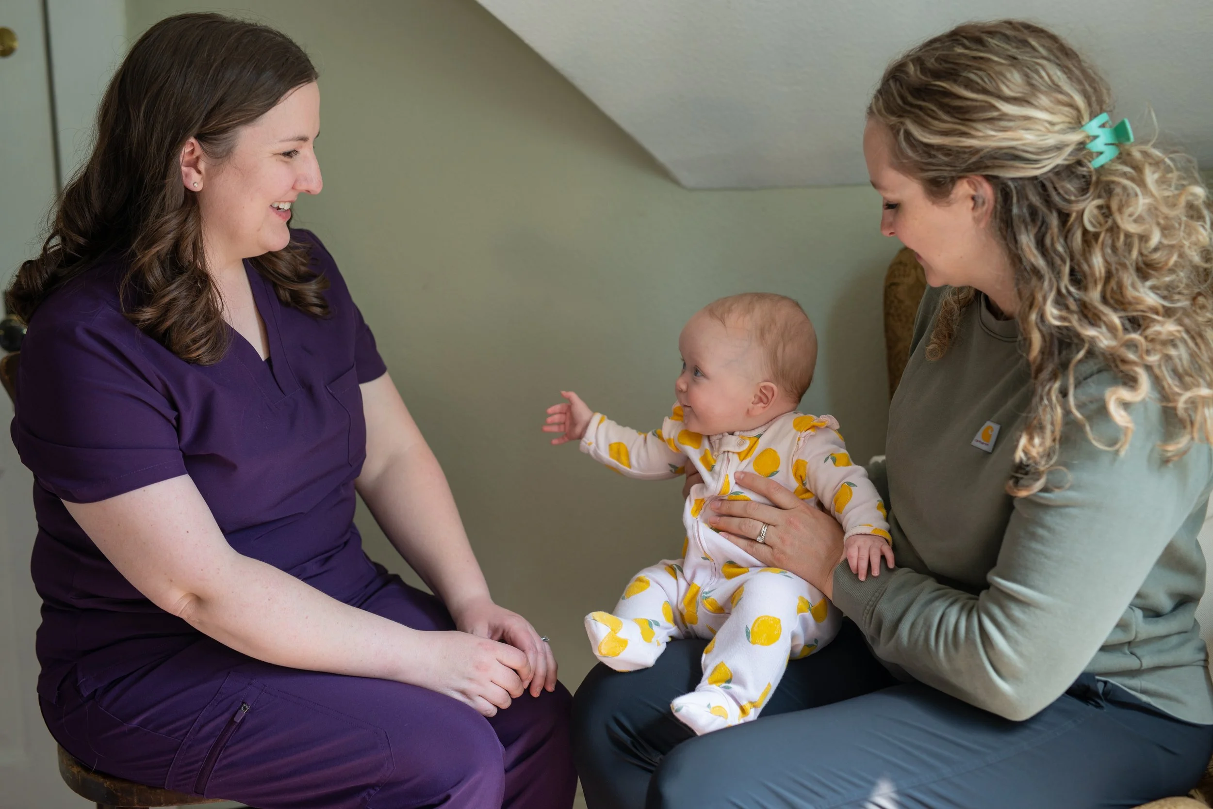 A nurse in purple scrubs sitting on a stool, a woman with long, curly blonde hair and a green shirt, and a baby dressed in yellow and white pajamas with a lemon pattern, engaging in a playful interaction in a healthcare setting.