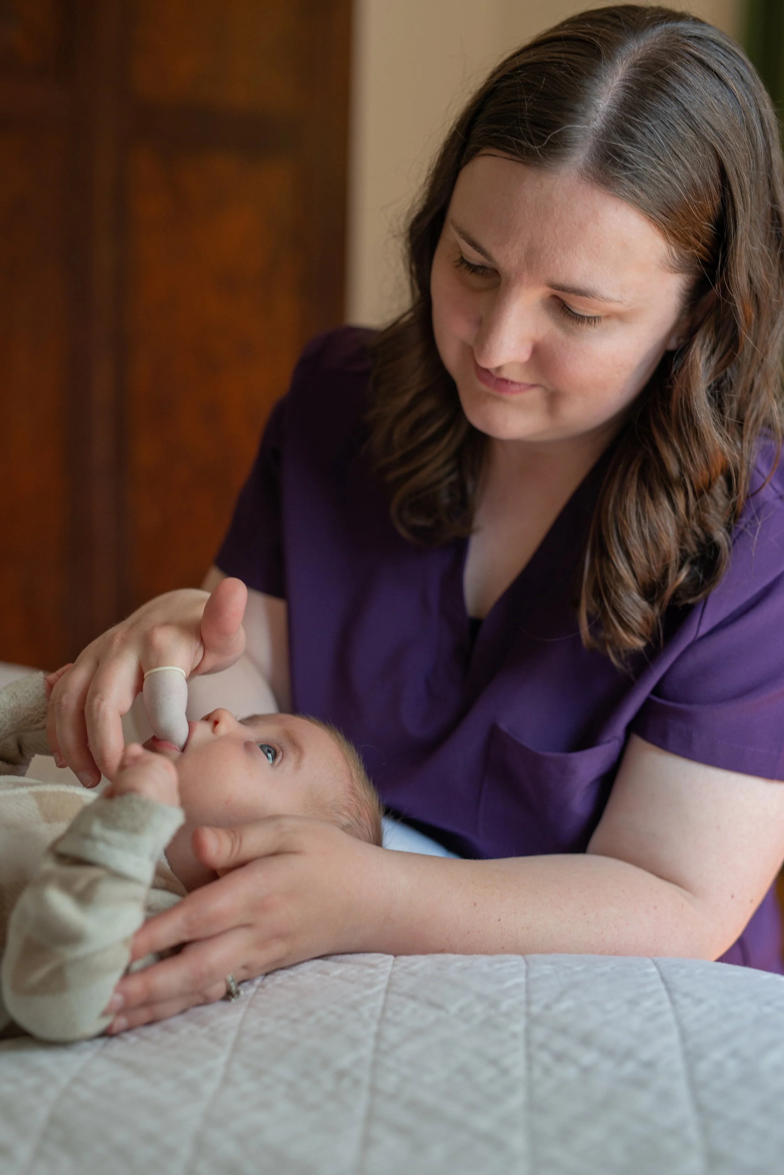 A woman in a purple shirt holding a baby with blue eyes and light hair, who is lying on a bed with a quilted beige blanket. The woman is smiling and looking down, while the baby gazes upward.