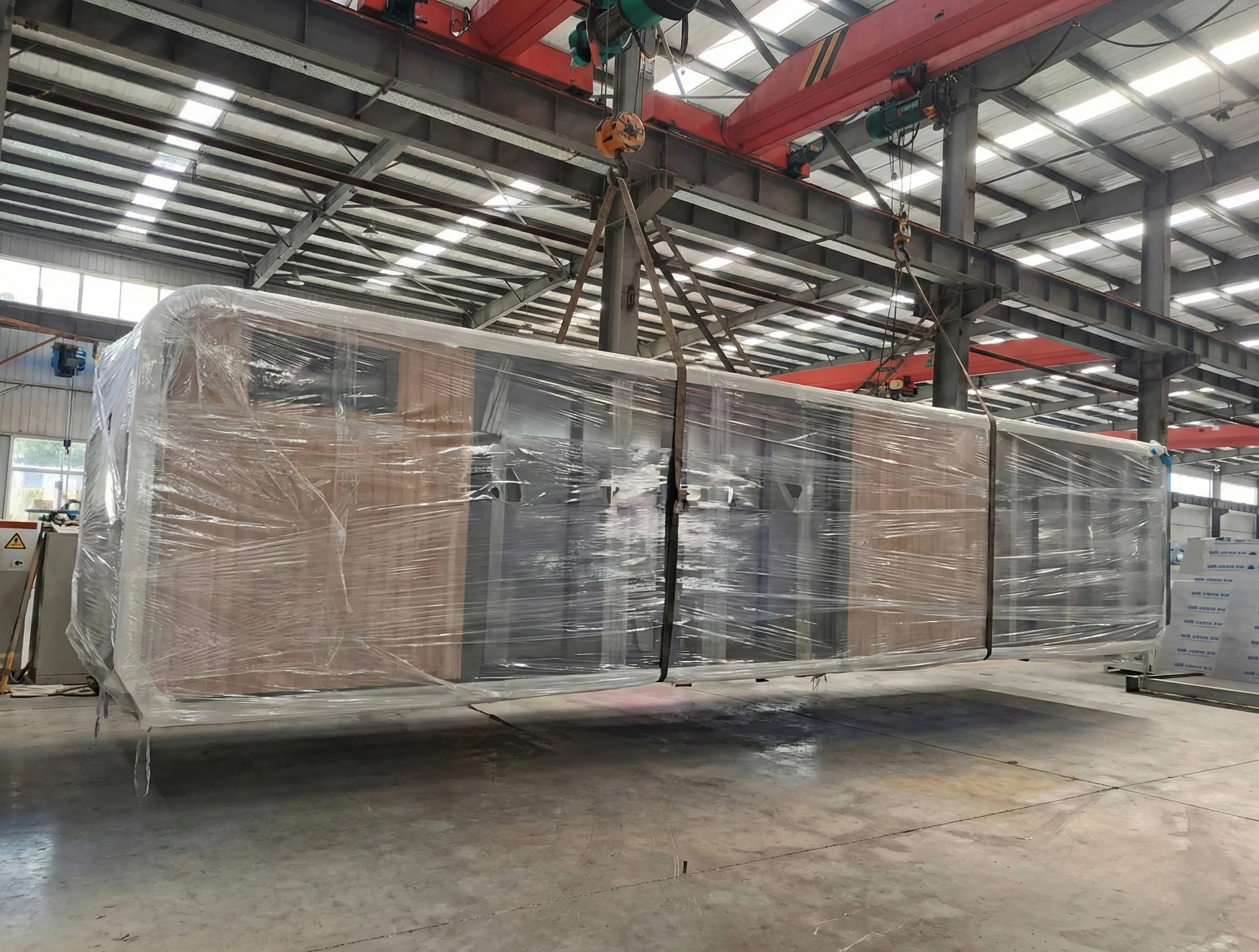 A large industrial crane suspends a wide, wrapped piece of machinery or equipment inside a warehouse with a metal roof.