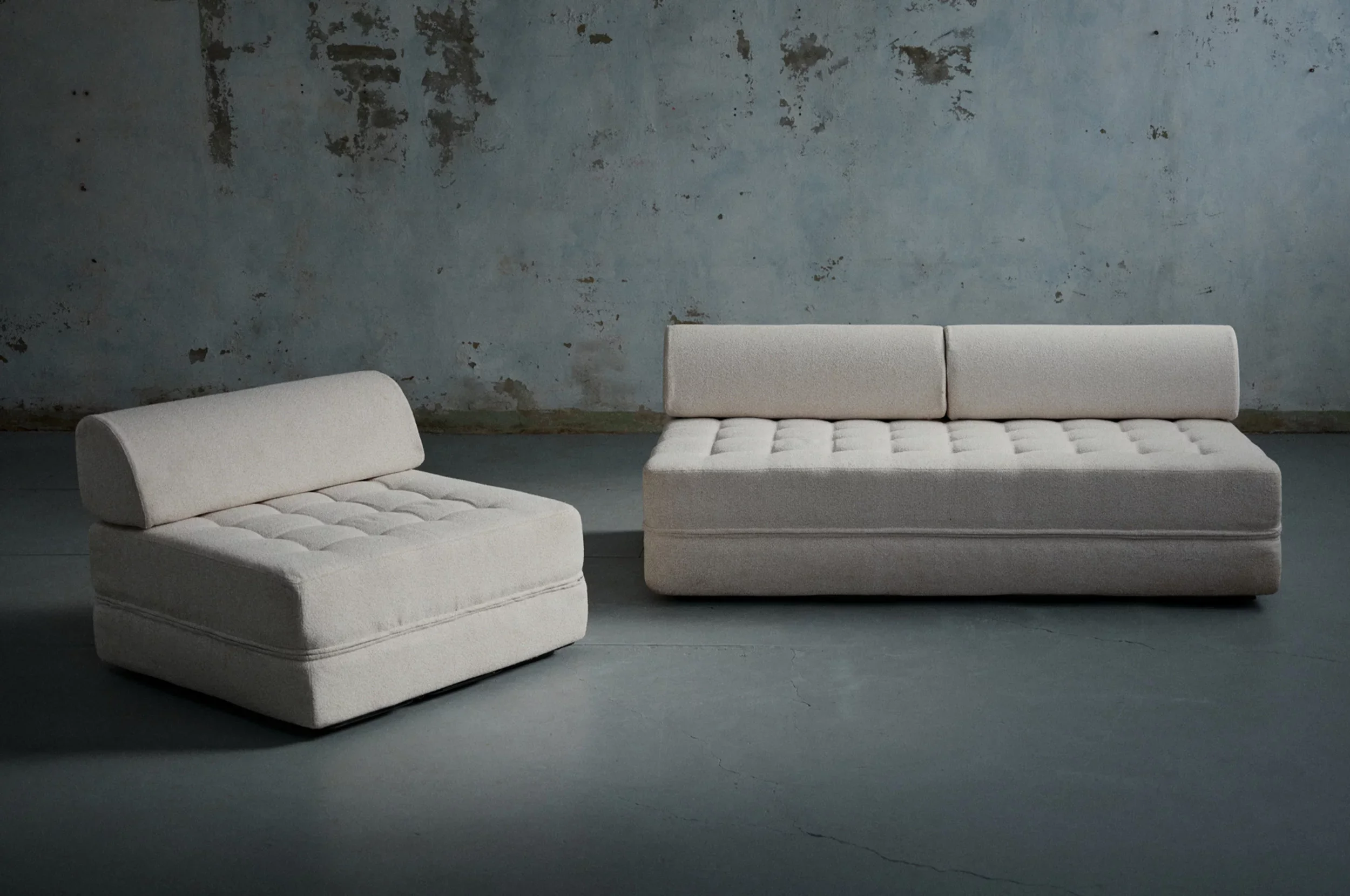 Two light-colored modern couches with tufted cushions in an industrial-style room with cracked concrete floor and distressed wall