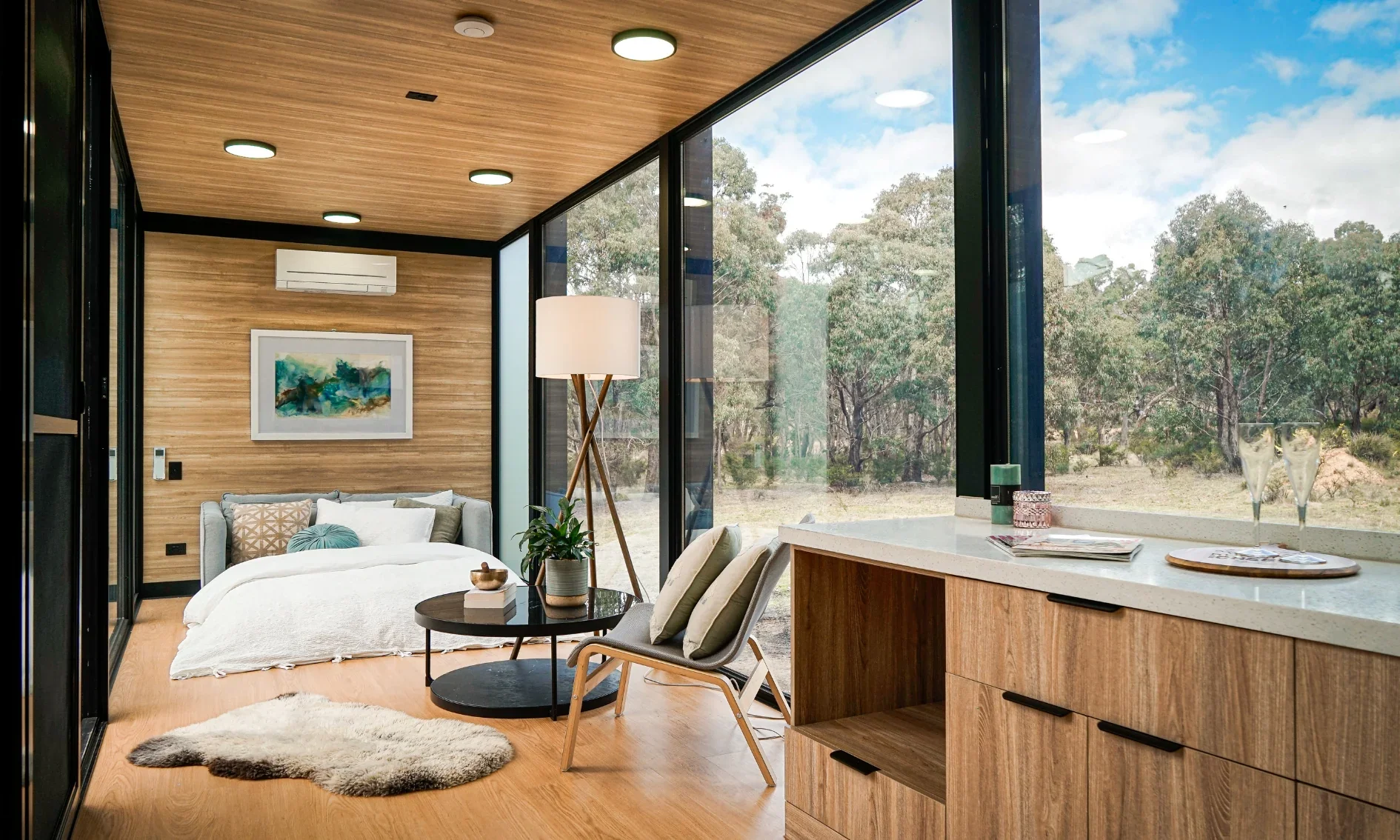Modern living space with a bed, a sofa, a round black coffee table, a floor lamp, and large glass windows overlooking trees.