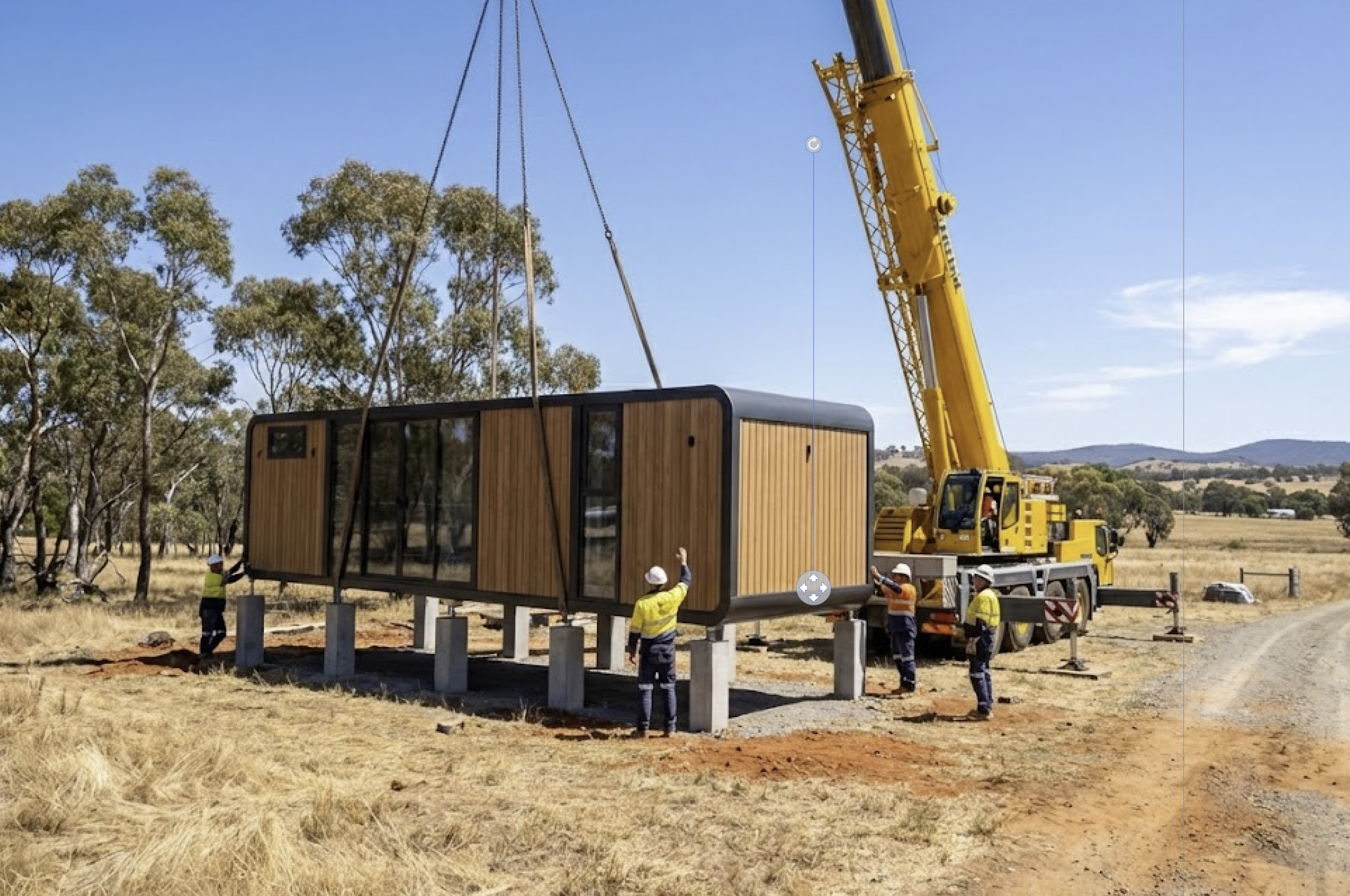 A construction site where a large yellow crane is lifting a prefab tiny house with wooden siding. Several construction workers in safety gear are guiding and observing the installation on concrete stilts in a rural area with dry grass, trees, and distant hills under a blue sky.